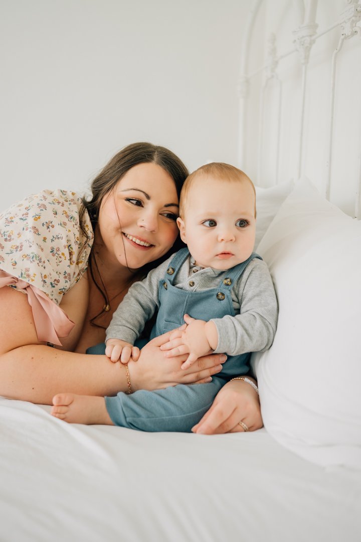  Parents sitting close with their baby in a bright Greenville natural light studio, soft neutral tones and joyful expressions.  Close-up profile of a one-year-old baby looking toward window light in a calm Greenville studio.  Father lifting his baby 