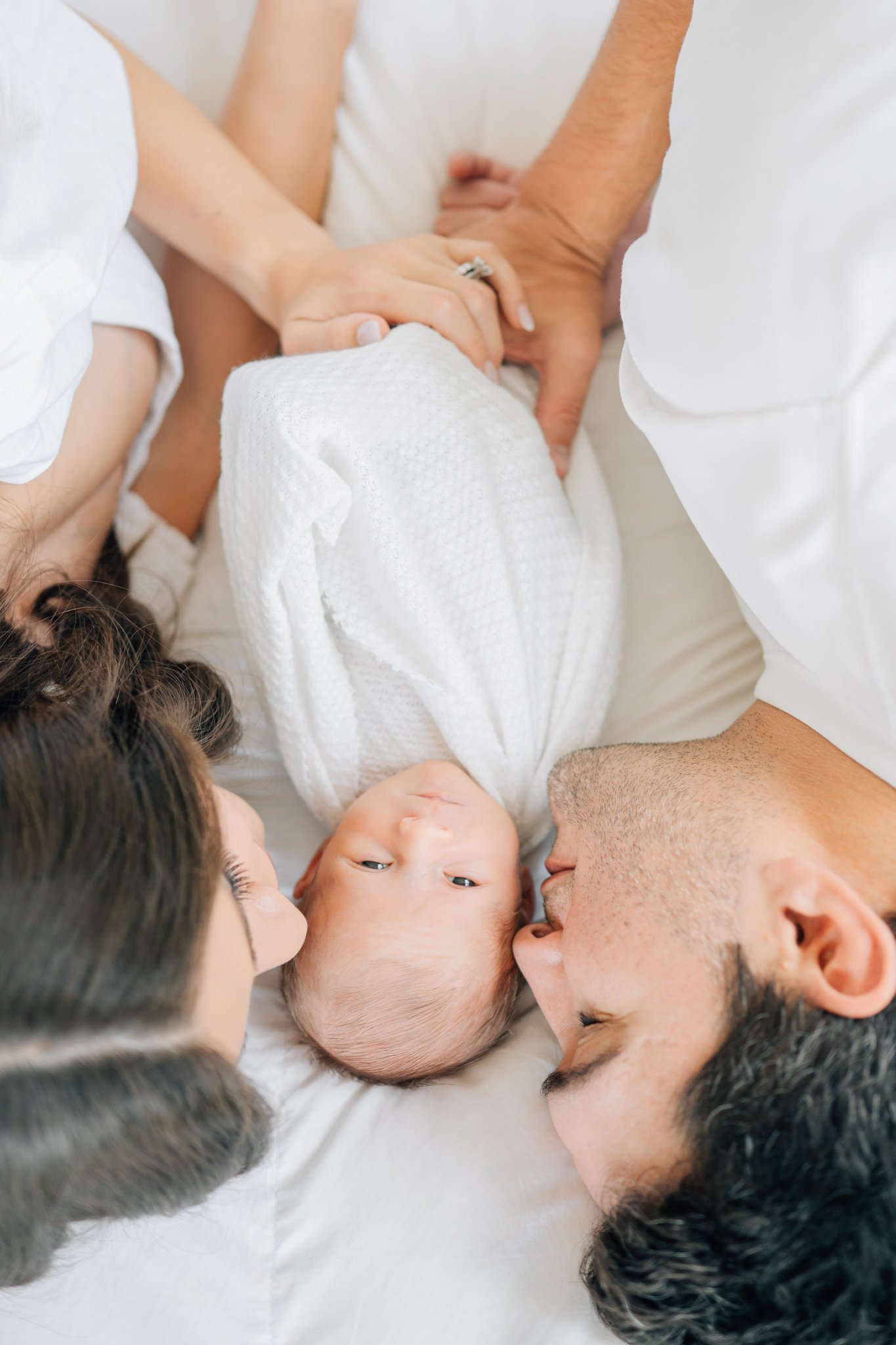 Newborn baby lying on a bed surrounded by parents and a woman, all looking at the baby and touching hands, in a white and soft-colored setting.