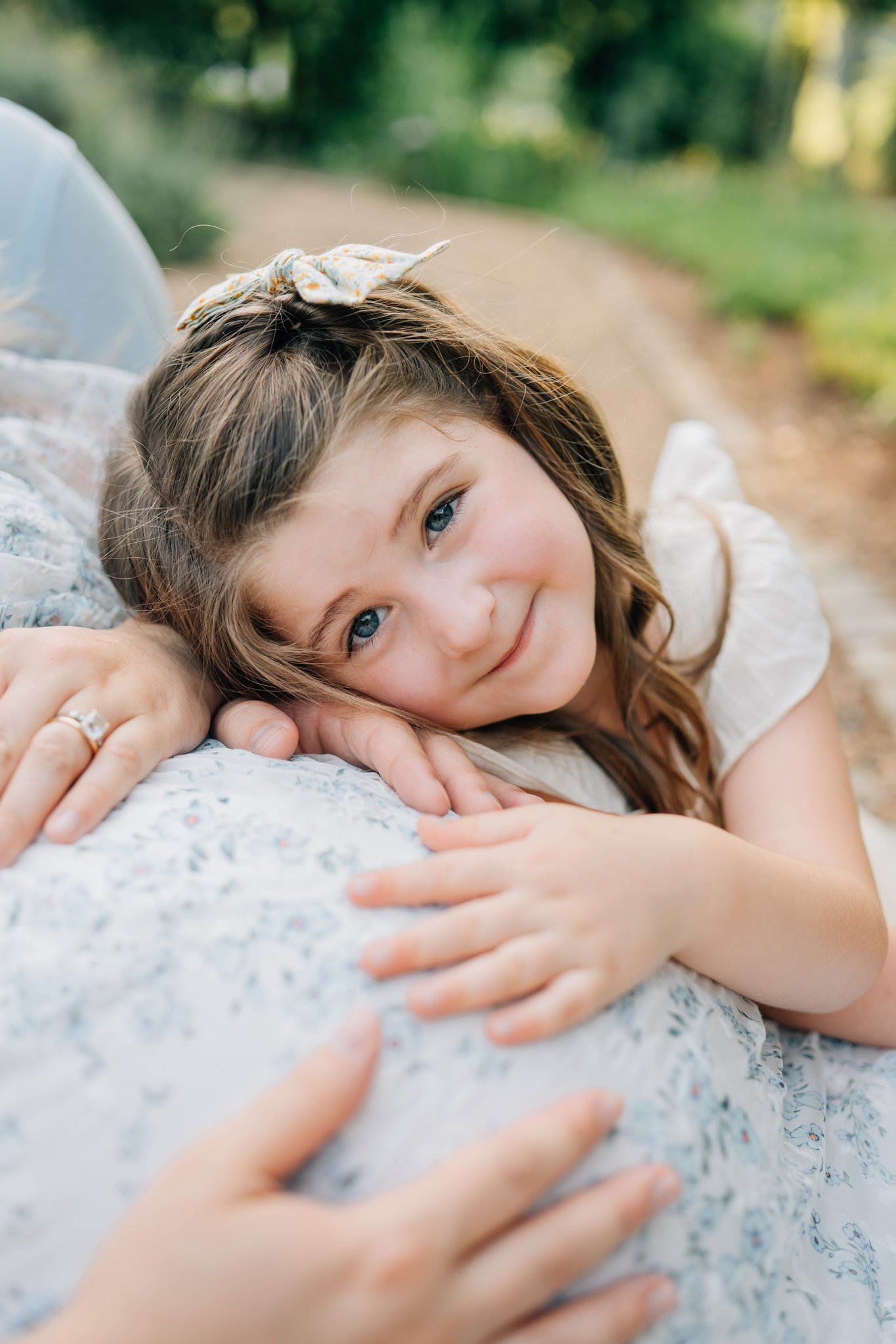 A young girl with blue eyes and brown hair, lying on her side outdoors, resting her head on someone's shoulder, with a gentle smile, during daytime.