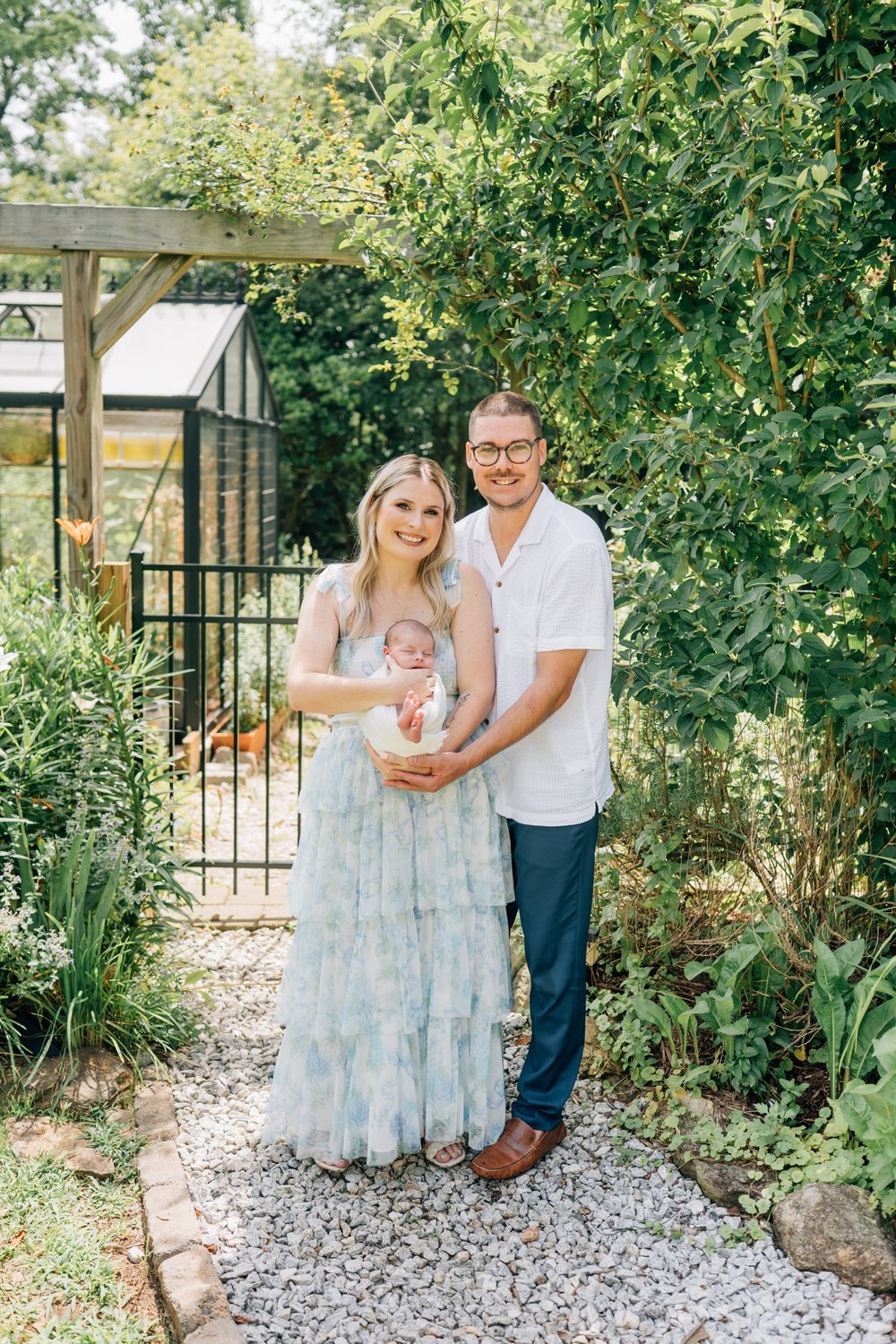 A happy couple holding their newborn baby in a garden setting surrounded by greenery, a greenhouse, and a black garden gate.