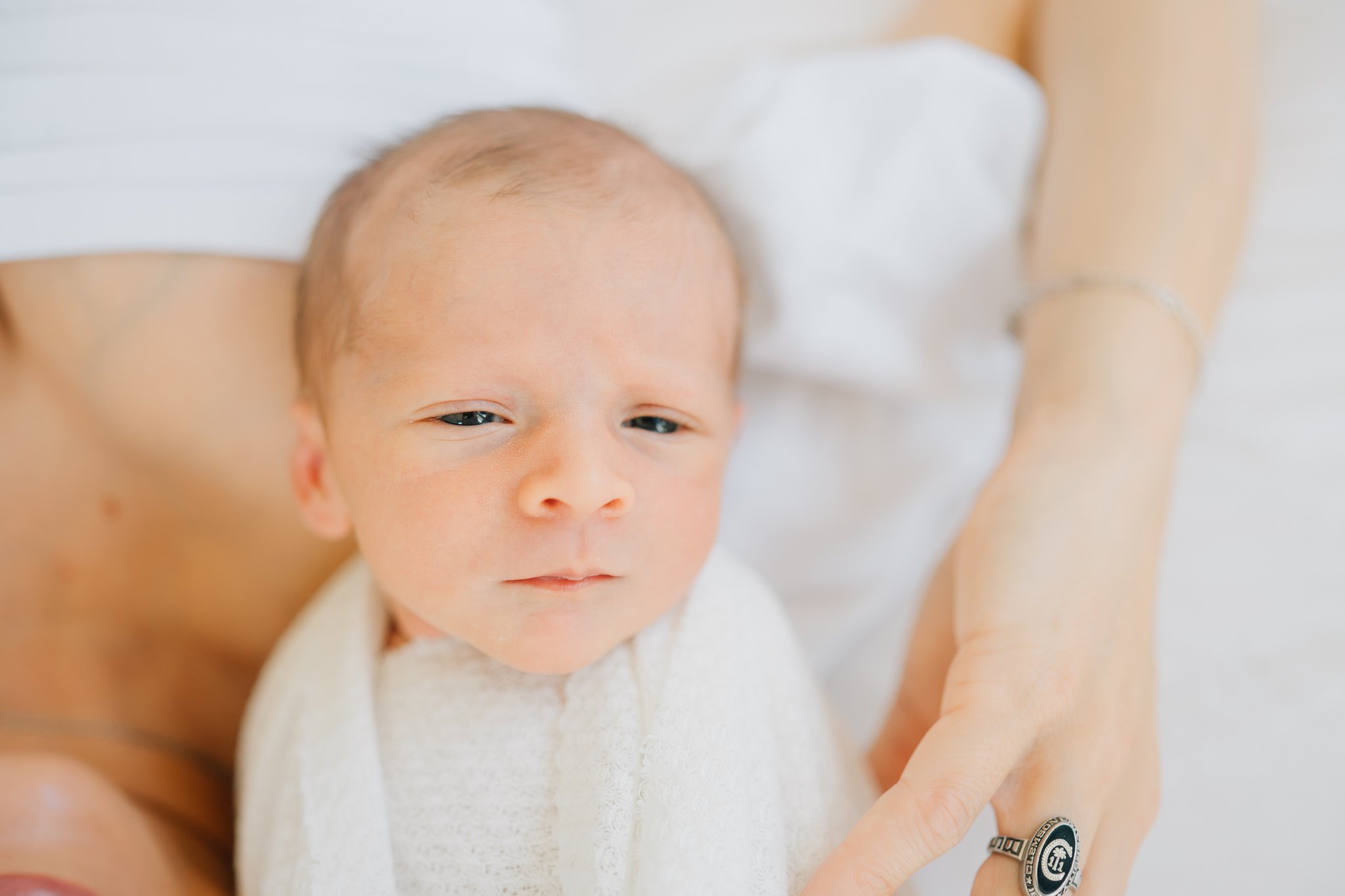Close-up of a newborn baby resting on an adult's chest, partially wrapped in a white blanket, with a blurred background.