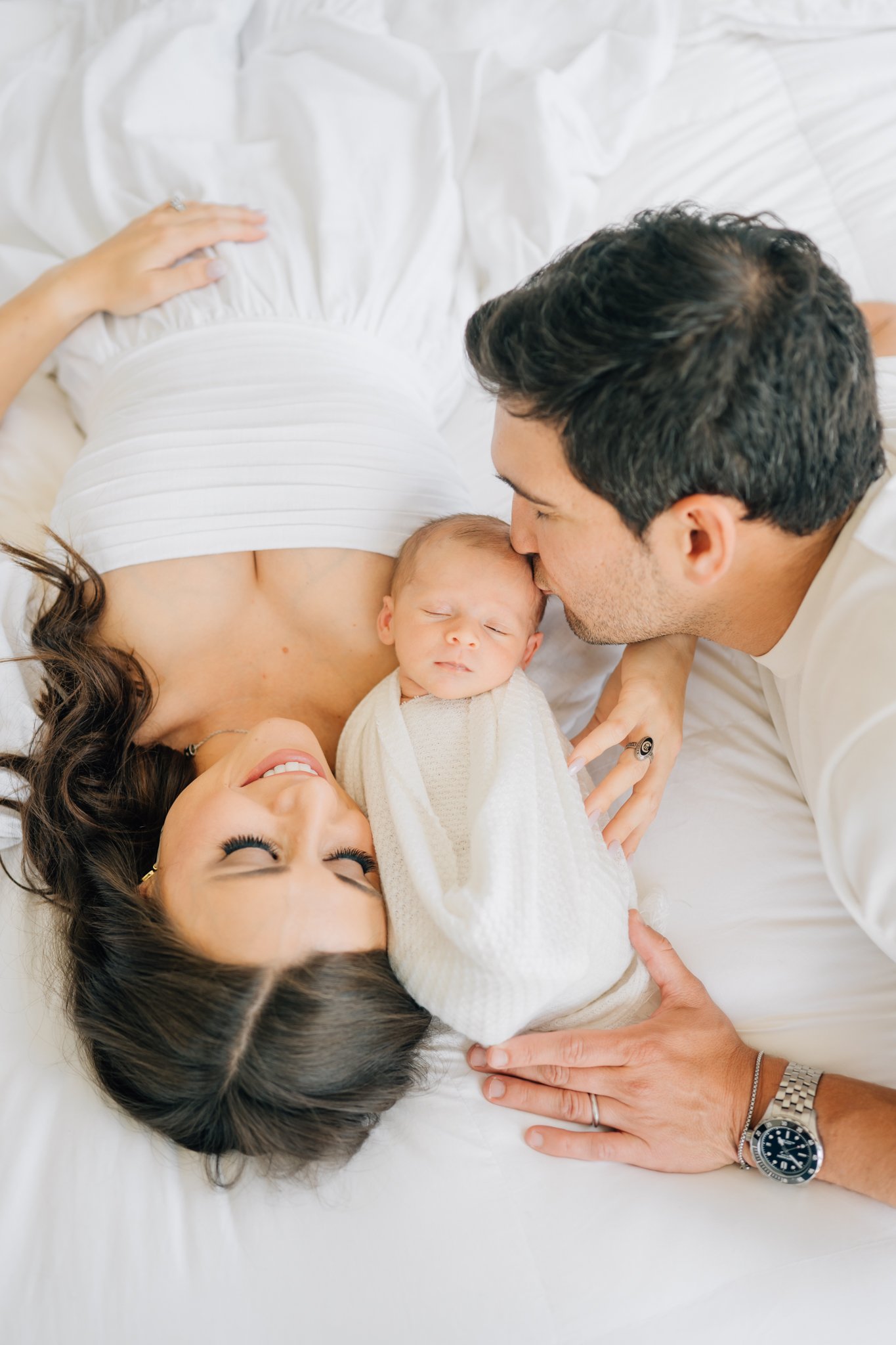 A happy family lying on a bed with a smiling mother, father kissing their newborn baby, who is wrapped in a white blanket.