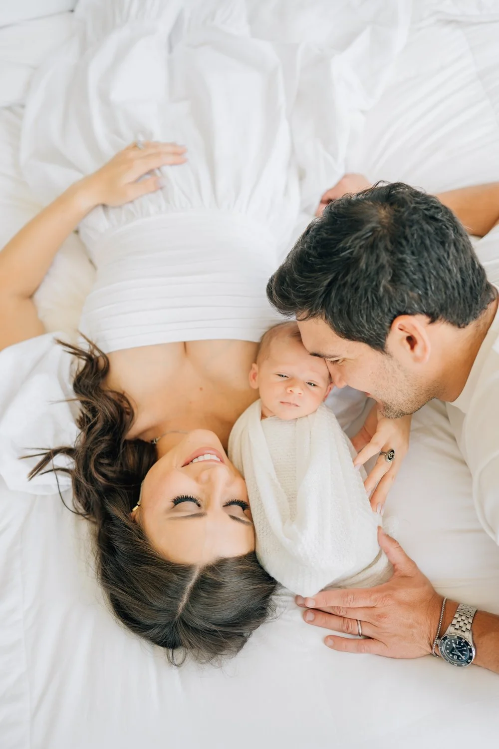Family lying on bed, mother pregnant, father, and newborn baby looking at each other.