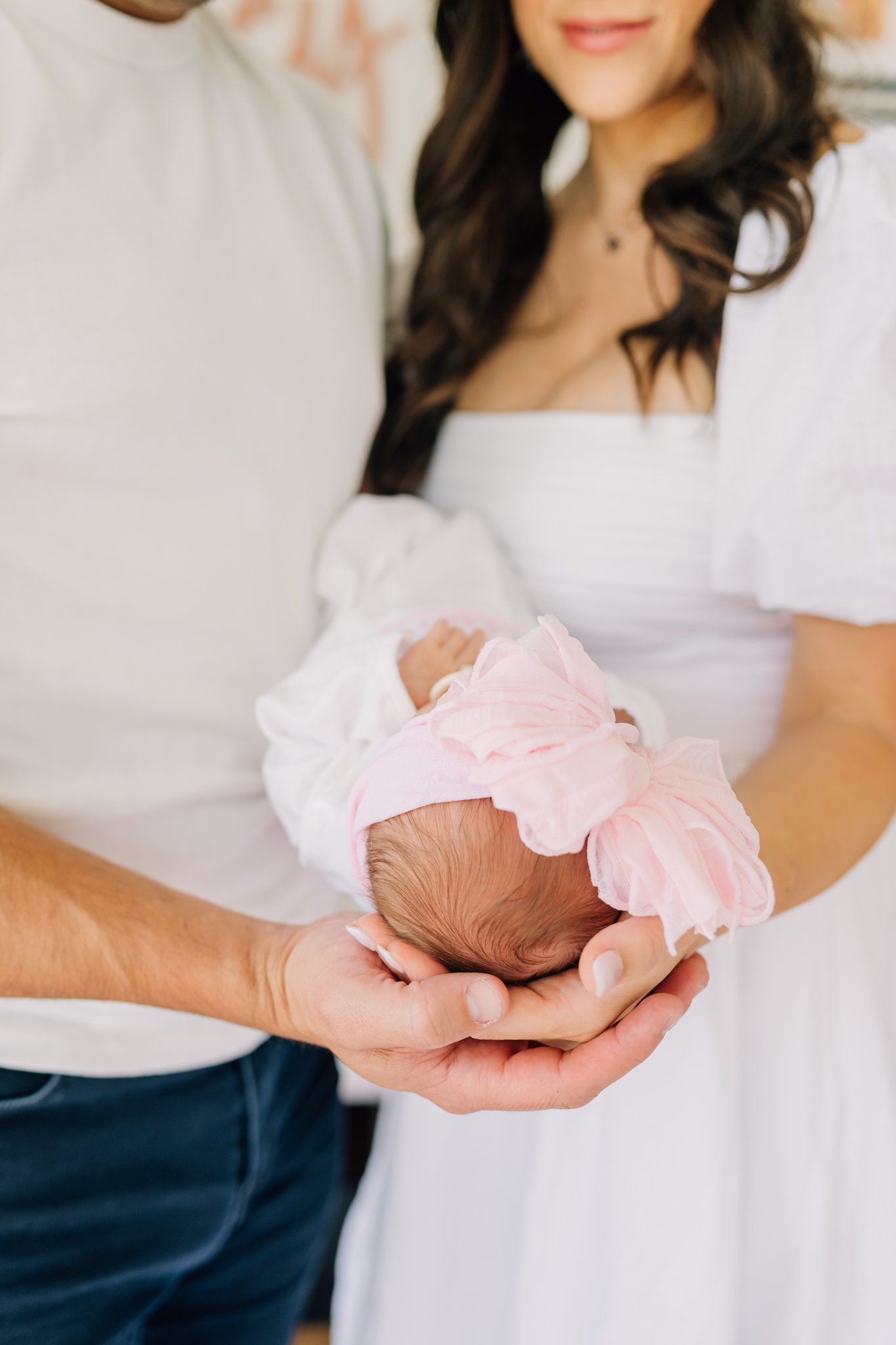 Close-up of a newborn baby girl wearing a pink headband, being held by a man and woman, with the woman smiling.