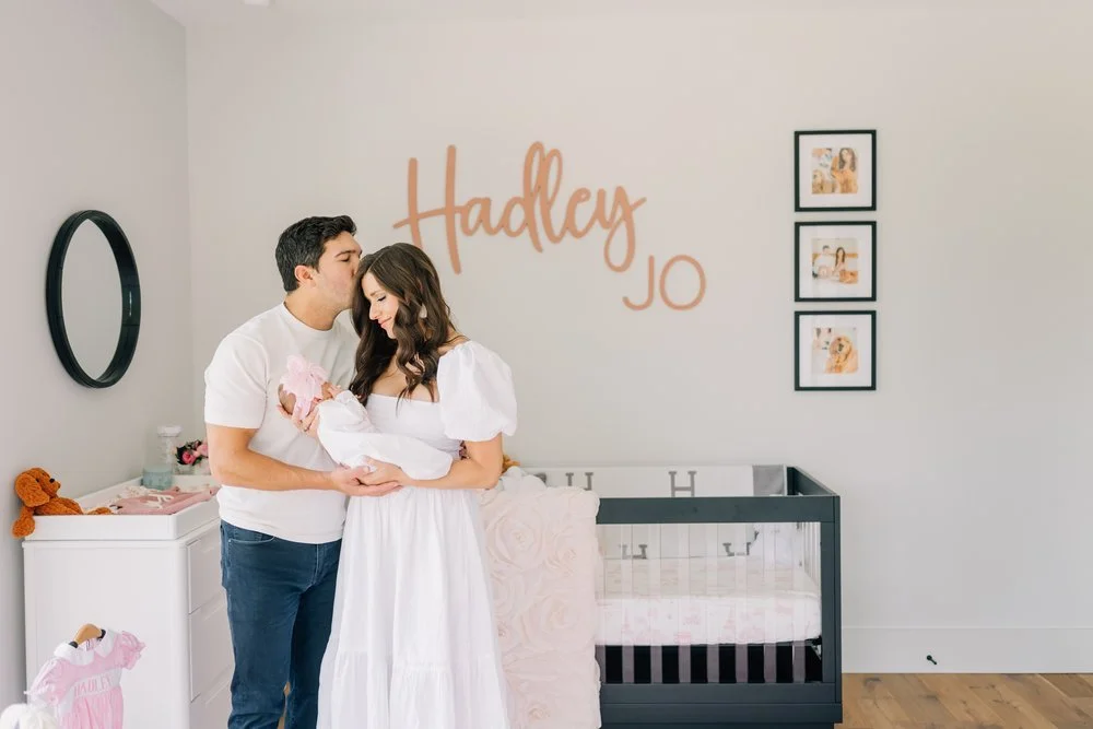 A couple holding a newborn baby girl in a nursery decorated with the names Hadley and Jo on the wall, framed photos, and a crib.