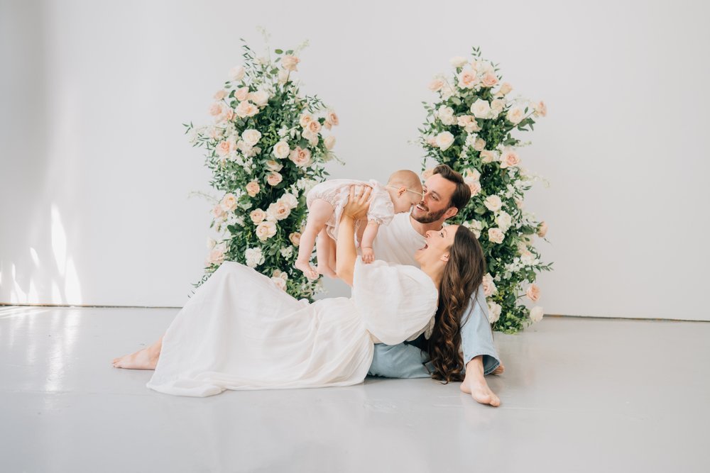 A joyful family of three with a woman, man, and baby, sitting on the floor in front of large floral arrangements of pink and white roses, smiling and playing together.
