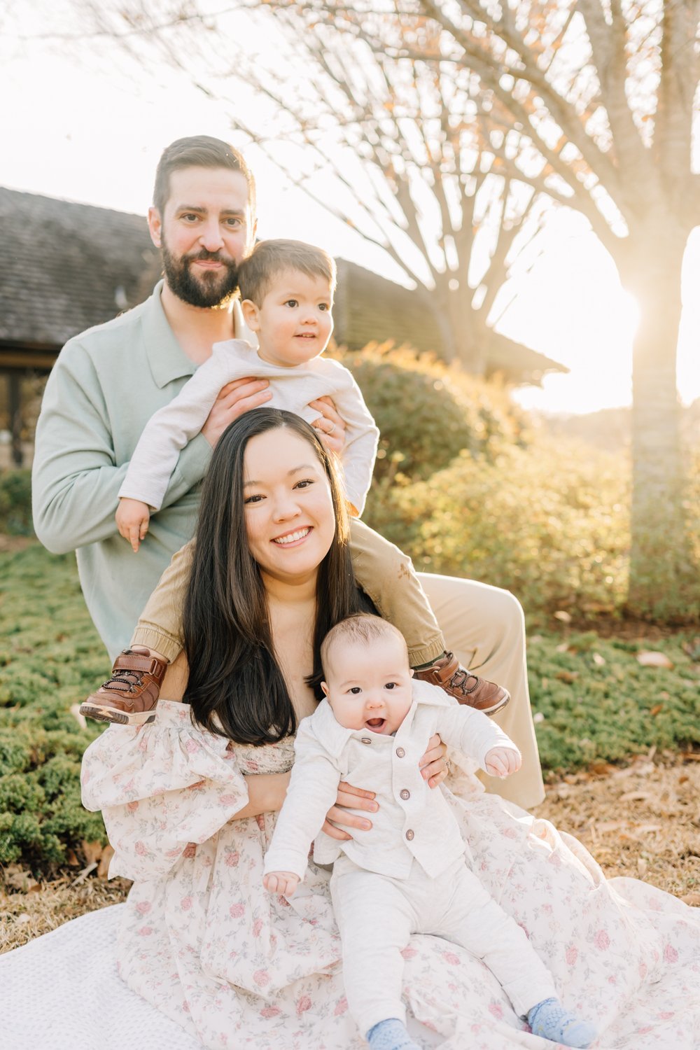 A family of four outdoors during sunset, with two adults and two children, smiling at the camera. The parents and children are sitting on a blanket on the grass, with trees and a house in the background.