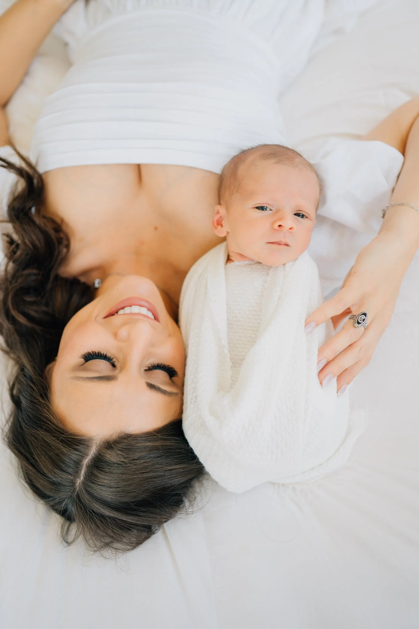 A woman lying on a bed with her eyes closed, smiling, and a newborn baby wrapped in a white blanket resting on her chest. The woman has dark hair and wears makeup, earrings, and a ring.