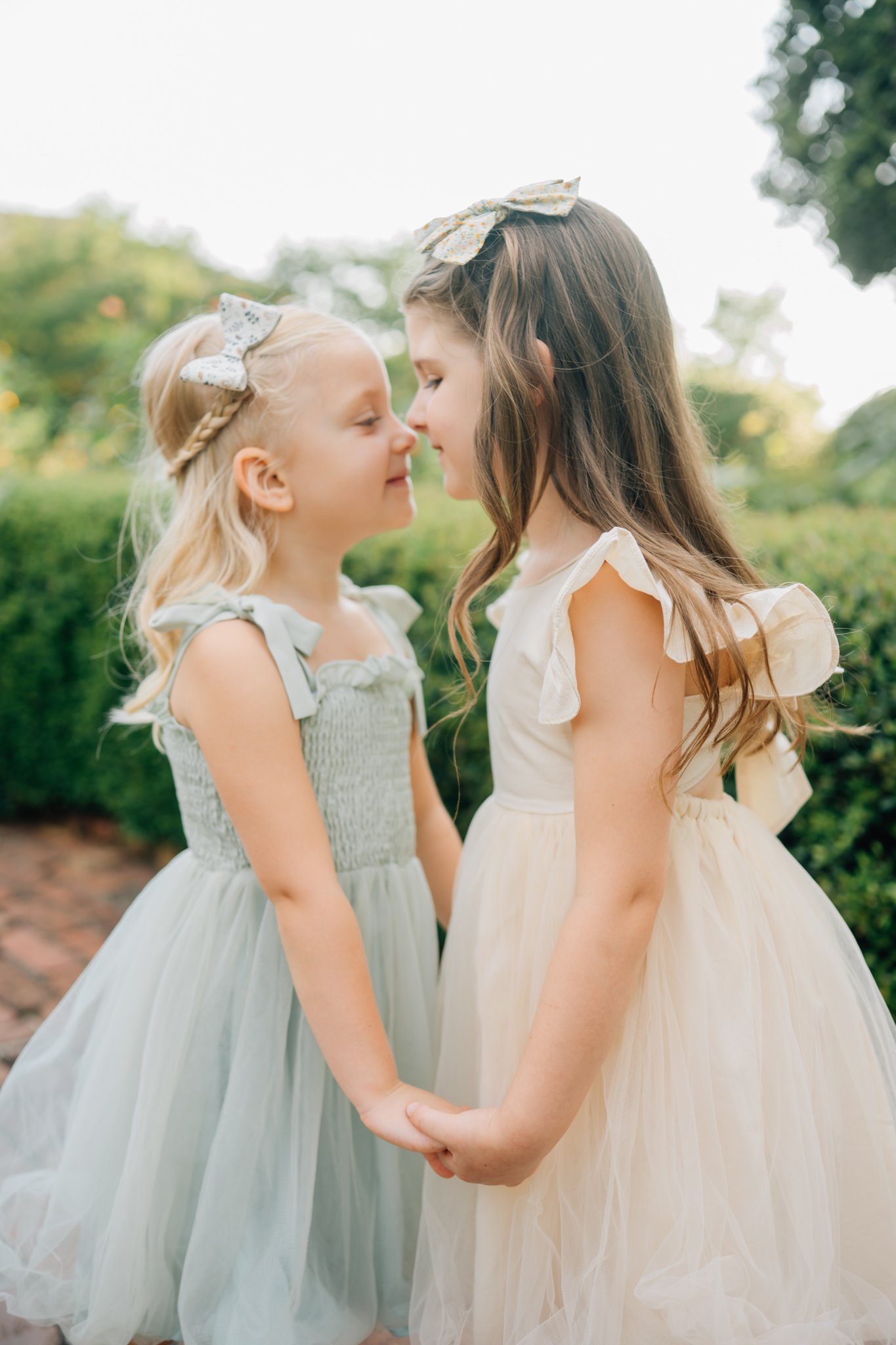 Two young girls in dresses holding hands and touching noses outdoors.