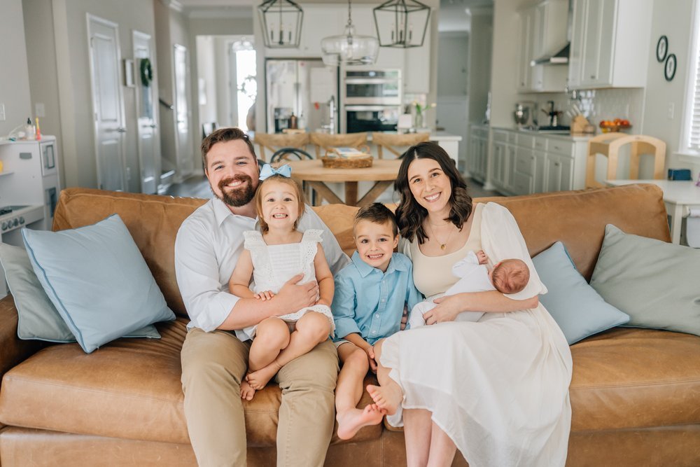 Family sitting on a sofa, smiling, with a woman breastfeeding a newborn, in a bright, modern kitchen.