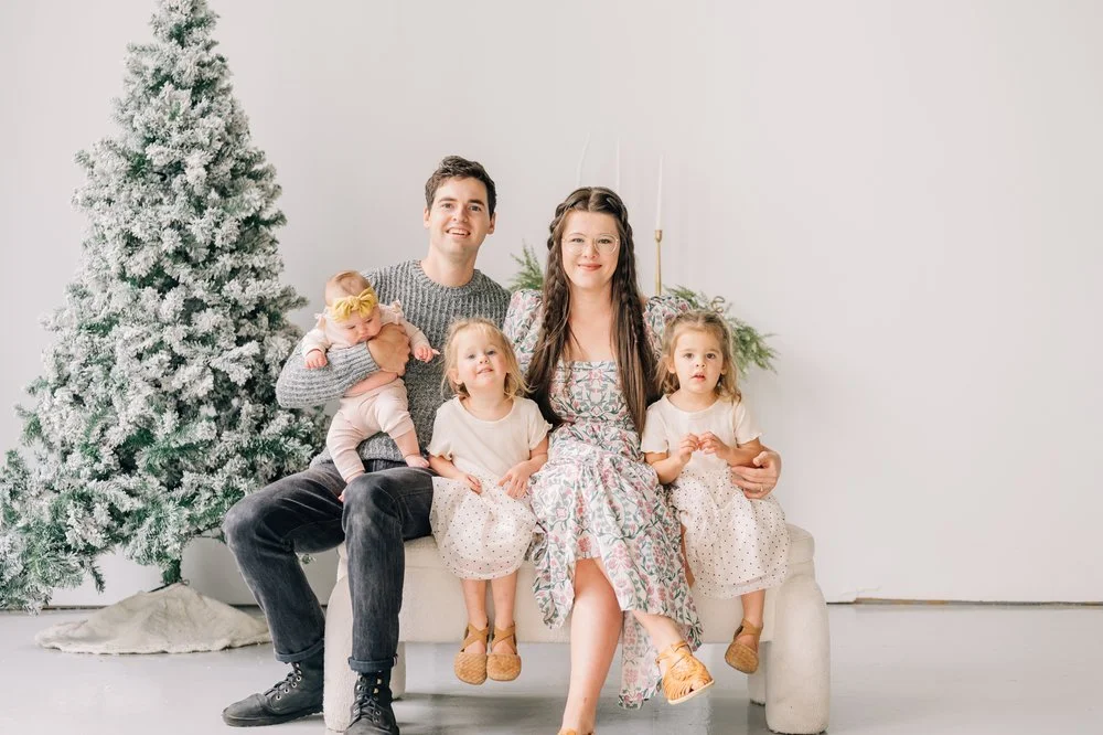 A family of five posing indoors next to a snow-covered Christmas tree. The father is holding a baby, while the mother sits beside two young children. They are all dressed in light, casual attire.