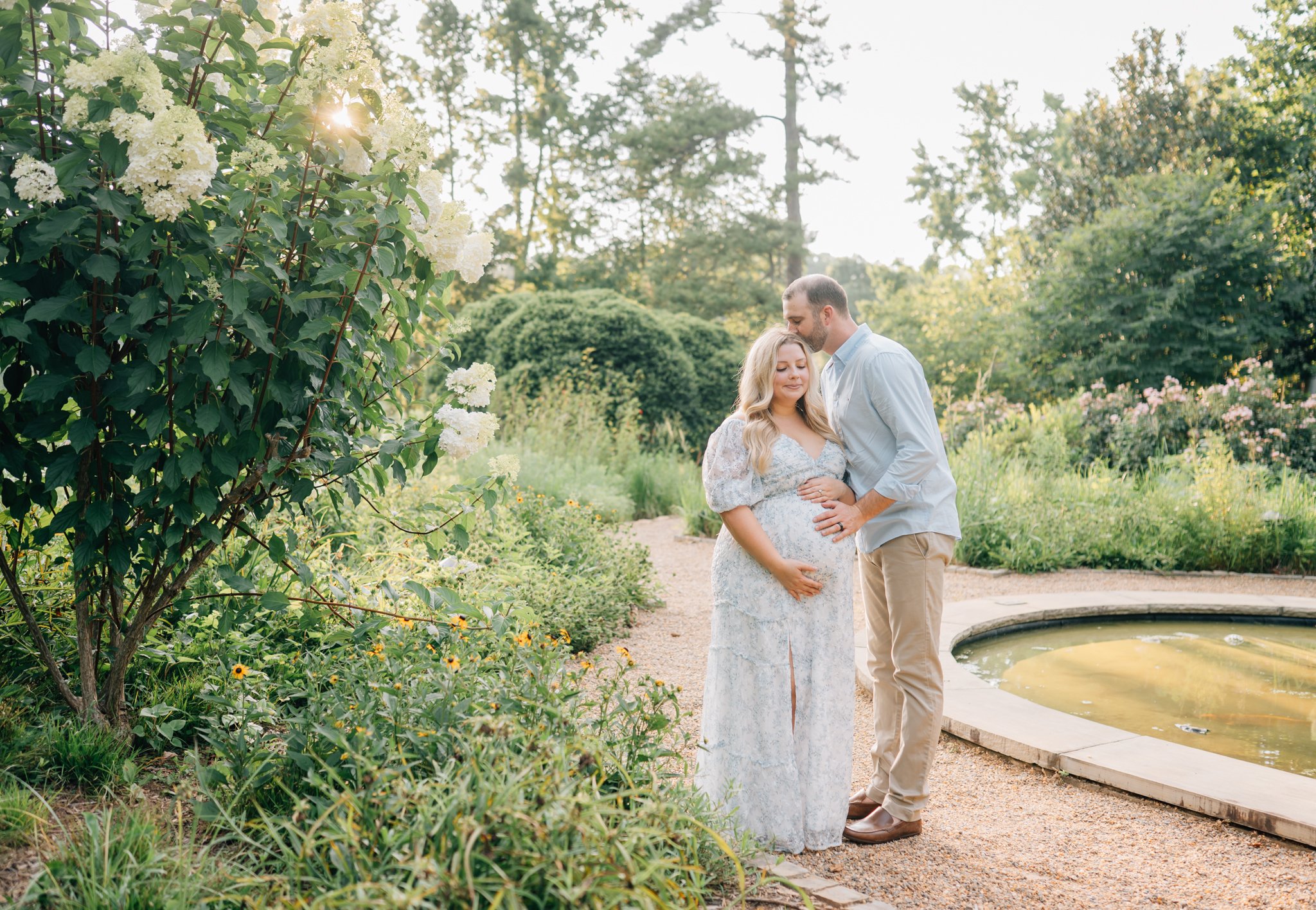 A pregnant woman and her partner standing in a lush, green garden with flowering bushes and trees, the woman cradling her belly and the man gently touching her stomach, with sunlight filtering through the trees.