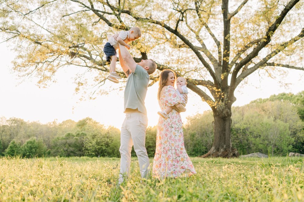 Family of four enjoying time outdoors in a grassy field during golden hour, with a large tree and green hills in the background. The father lifts a young boy in the air, while the mother holds a baby girl.