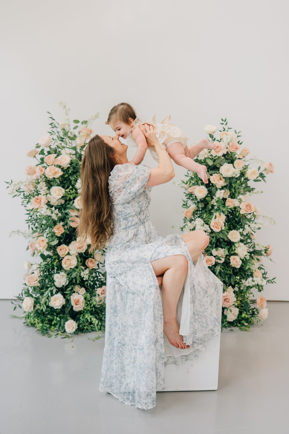 A woman holding a young girl in the air, both smiling and touching noses, with a backdrop of pink and white roses and green foliage.