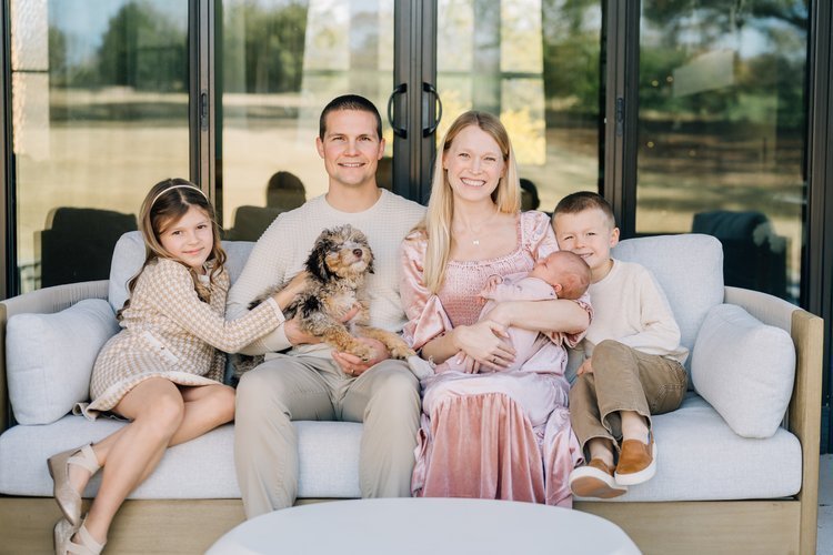 Family of five sitting on a sofa with their dog, smiling. The family includes a young girl, a man, a woman holding a baby, and a young boy, with a glass porch door behind them.