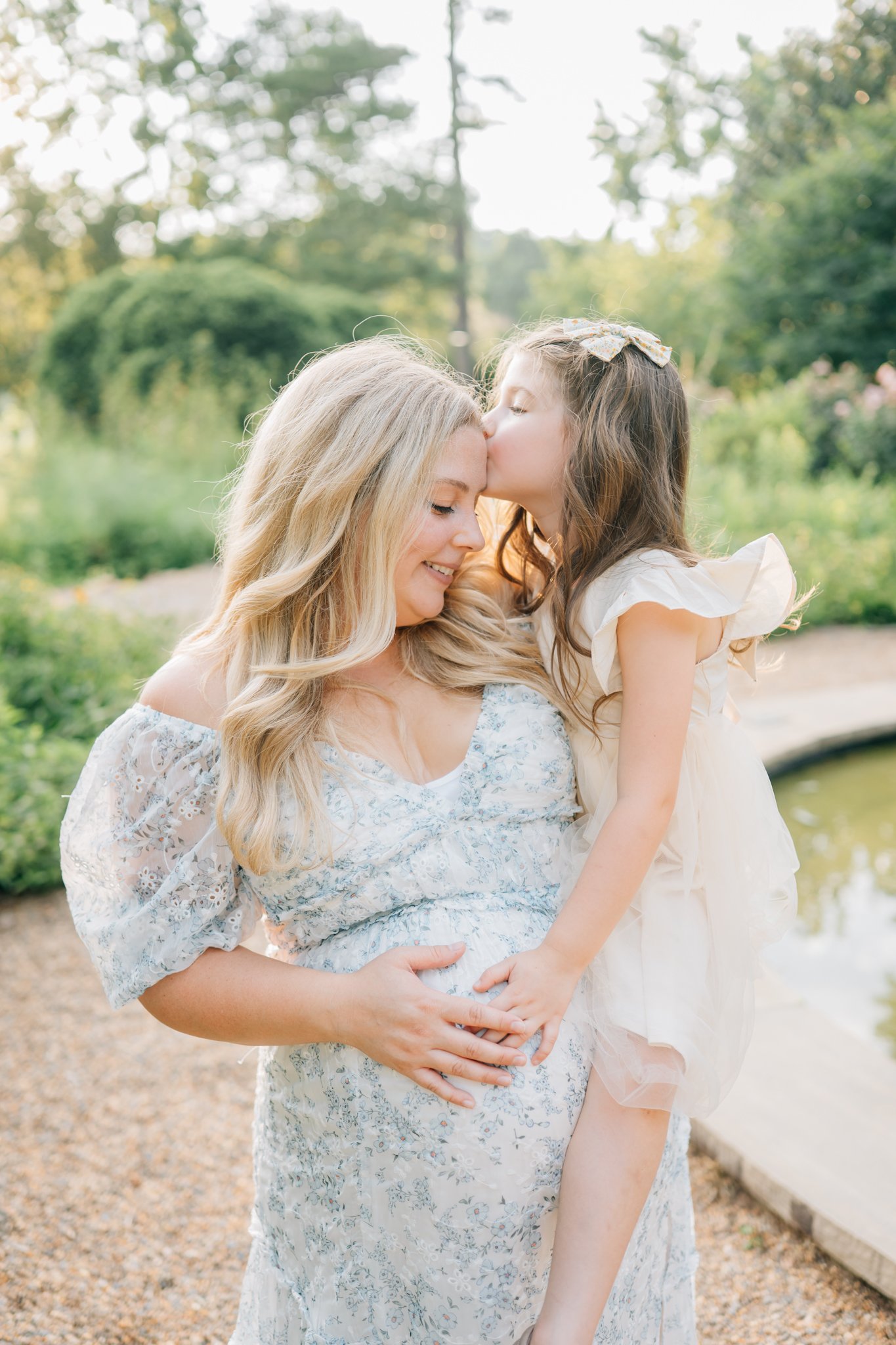 A woman holding a young girl outside near a pond, the girl is giving the woman a kiss on the forehead. The setting is a lush, green garden during daylight.