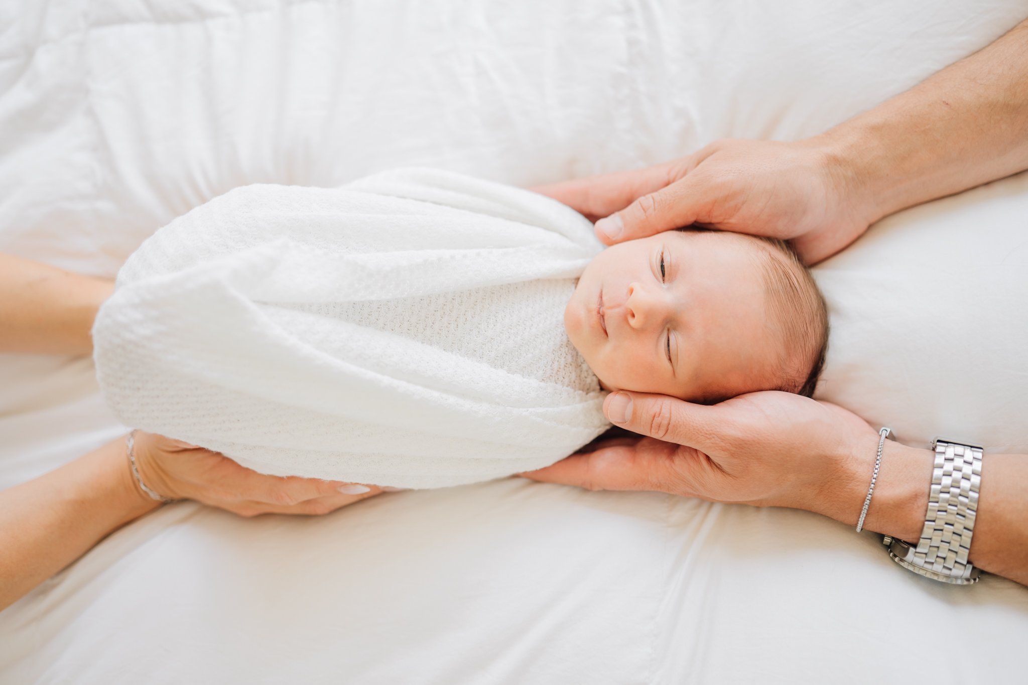 Close-up of a newborn baby sleeping, gently cradled by adult hands, wrapped in a white blanket.