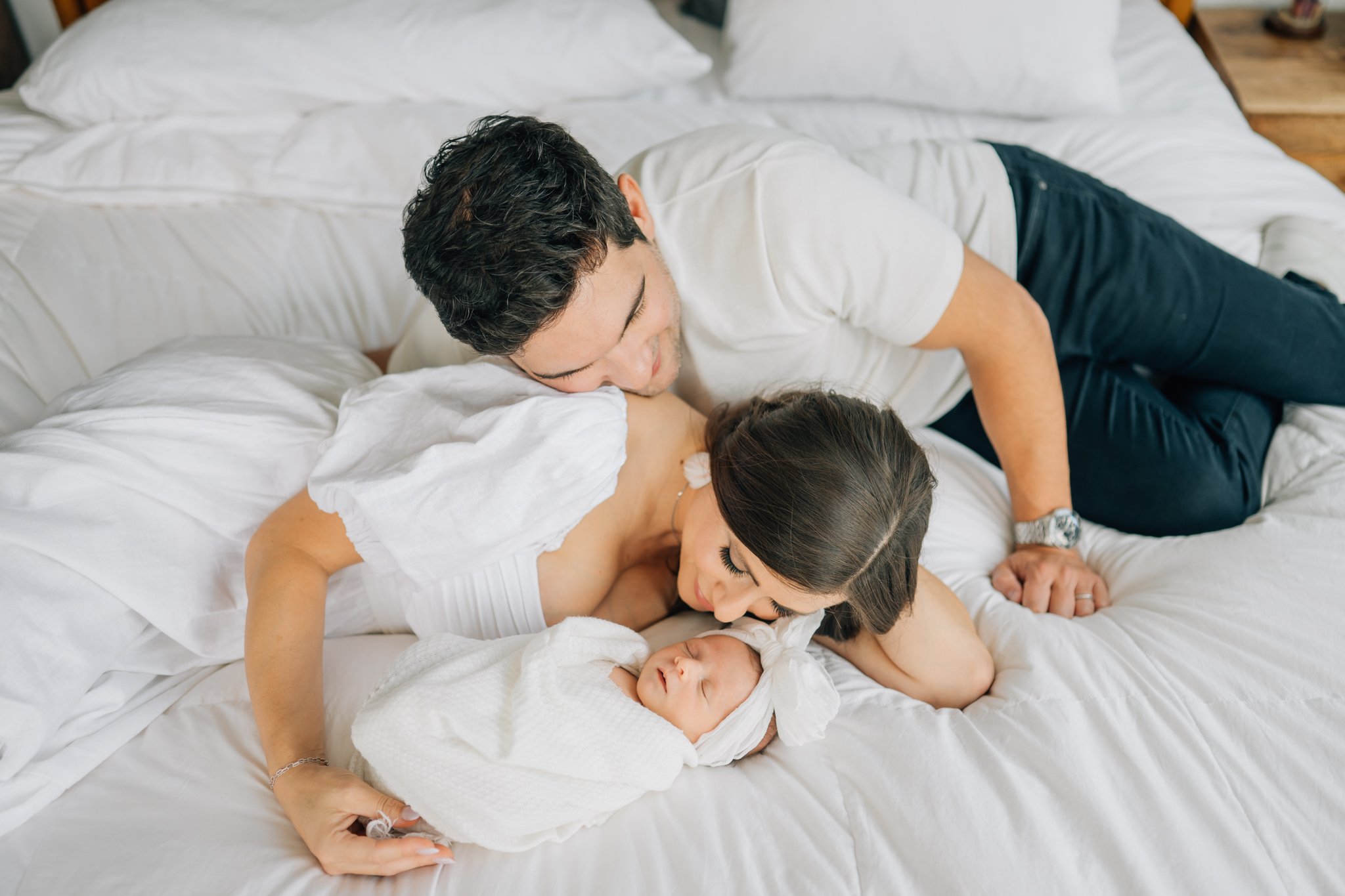 A couple lovingly gazes at their sleeping newborn baby wrapped in a white blanket on a bed with white sheets, with the mother lying on her side and the father leaning over her.