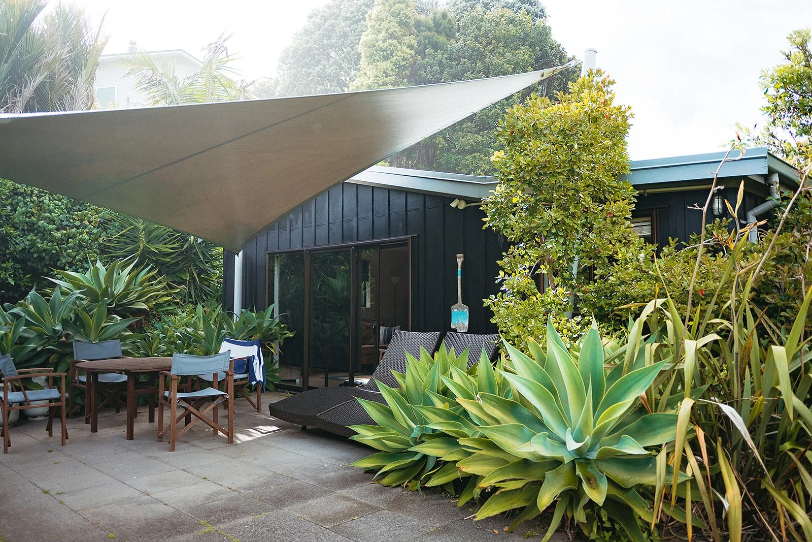 Architectural exterior photography of a modern dark-stained building with a dramatic shade sail canopy, outdoor lounge furniture, and lush agave and tropical plantings. Ideal for architectural firms, real estate brands, outdoor living product campaig