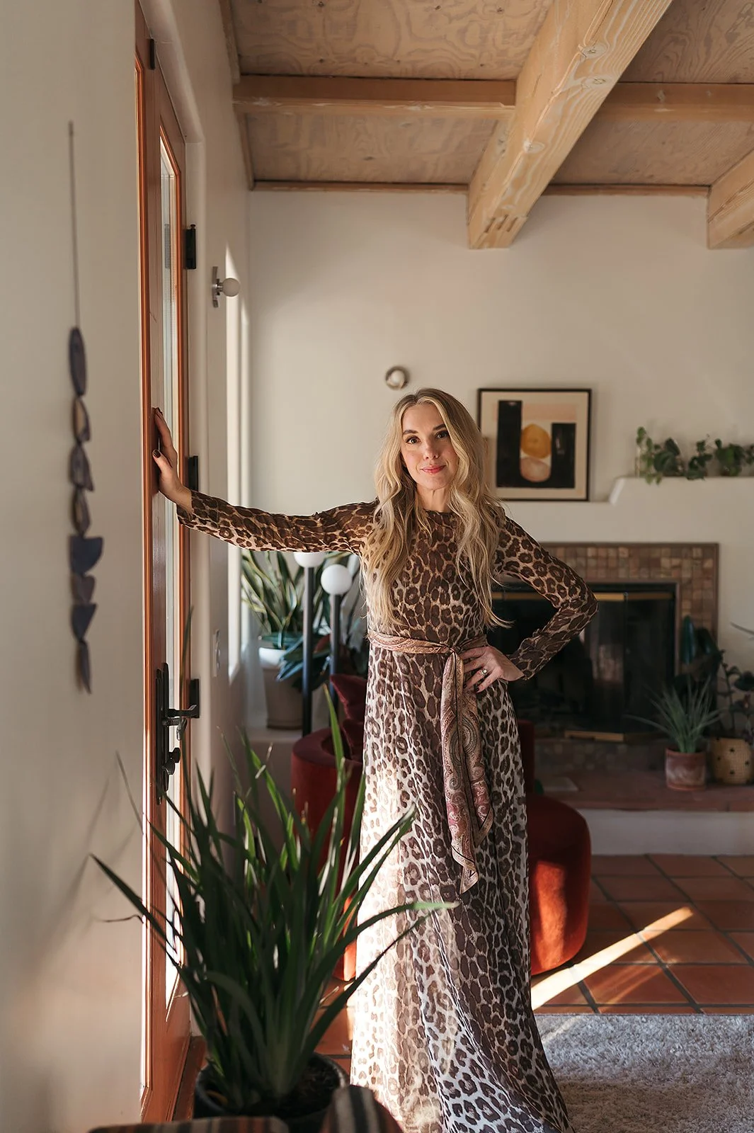 Fashion and lifestyle portrait of a woman in a flowing leopard print maxi dress, posing in a sun-drenched bohemian interior with exposed wood beam ceilings, indoor plants, and curated art. Perfect for fashion brand campaigns, clothing lookbooks, inte