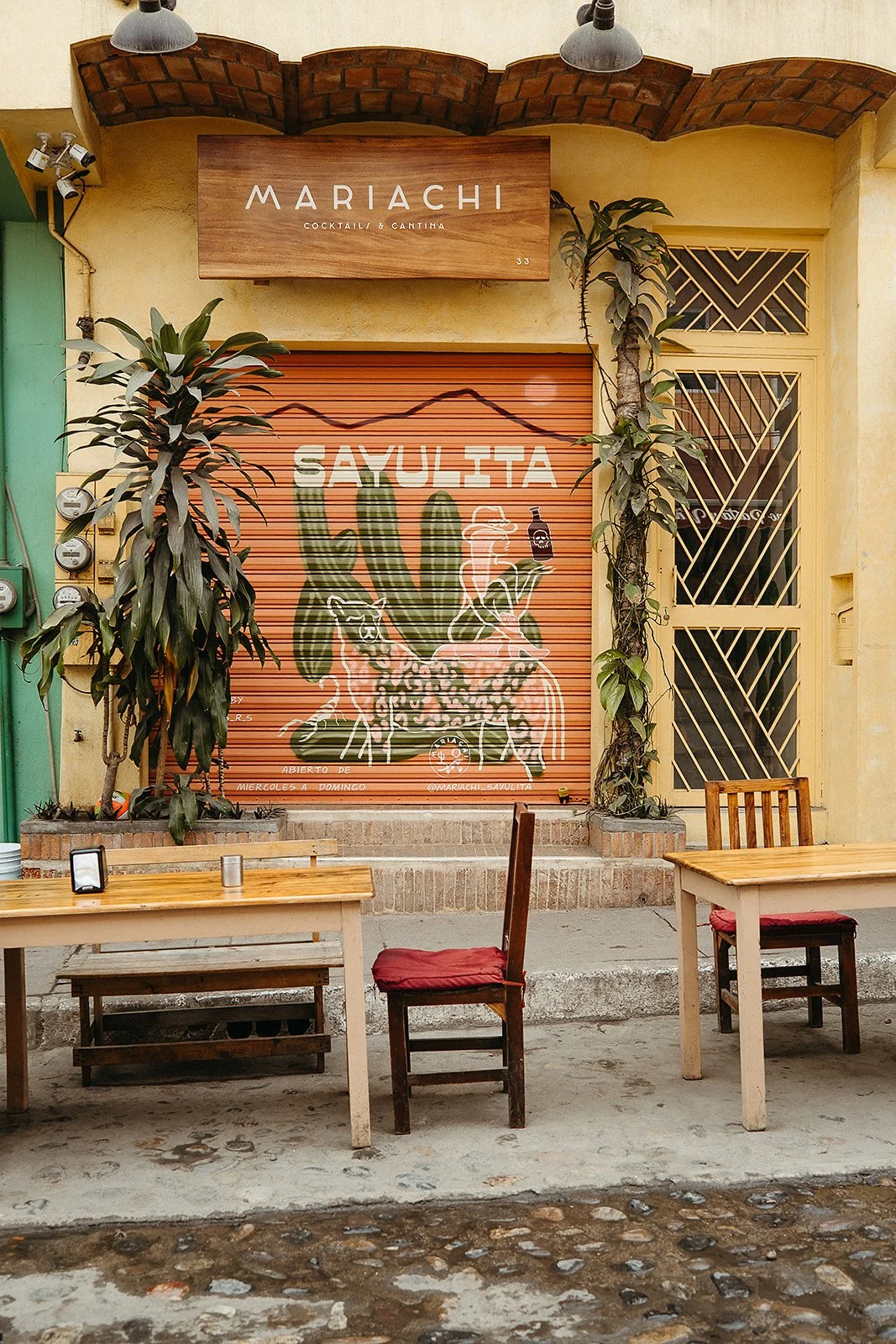 Vibrant street photography in Sayulita, Mexico, featuring a painted storefront shutter with cactus artwork, rustic wooden café tables on cobblestone streets. Colorful travel and destination photography perfect for tourism brands, Latin American trave