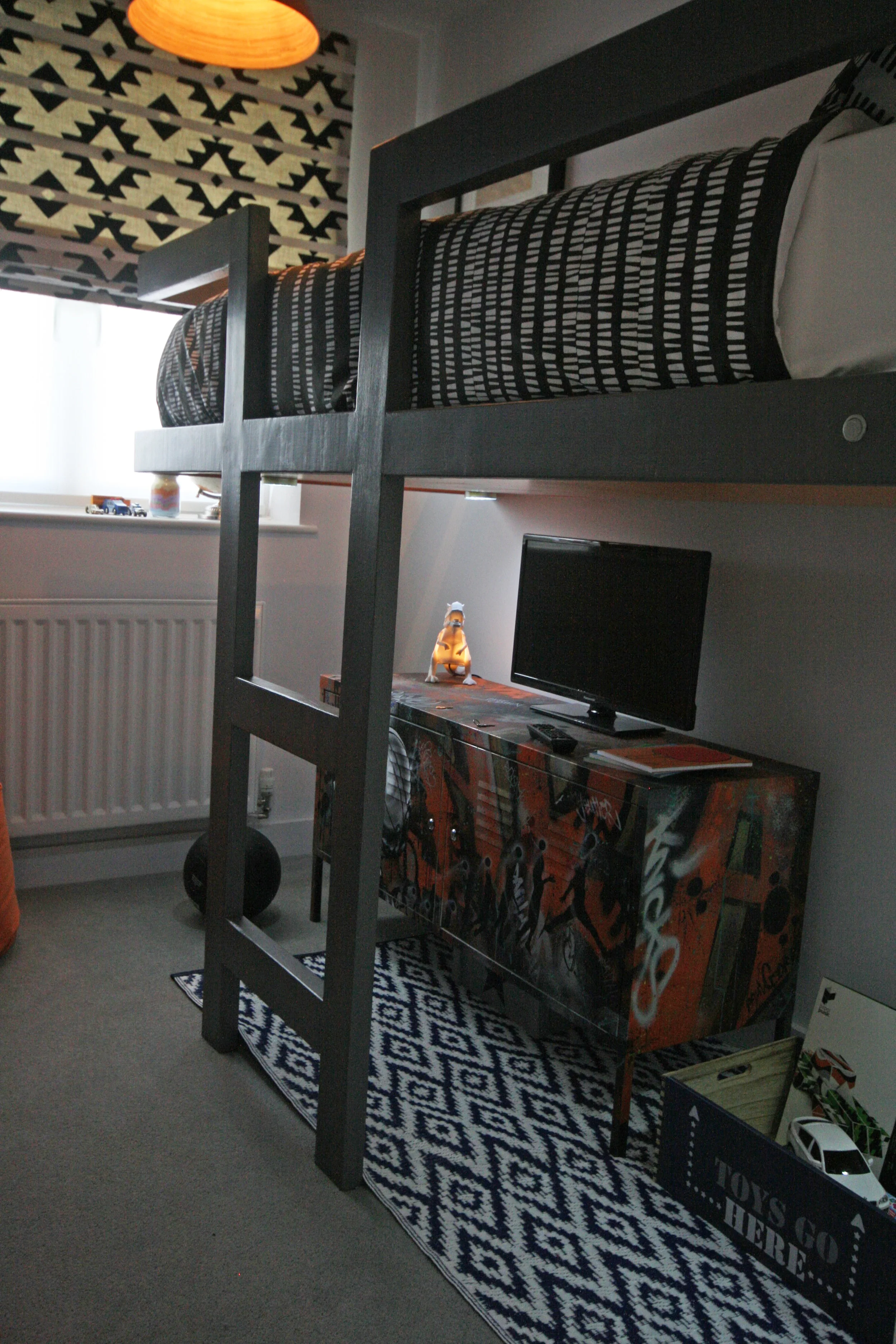Charcoal high level, built in bed with geometric soft furnishings and a graffitied cabinet.