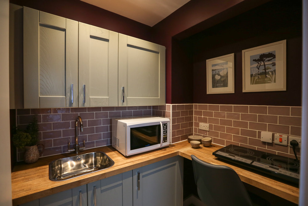 Plum coloured kitchenette with lilac coloured tiles and sage kitchen units. 