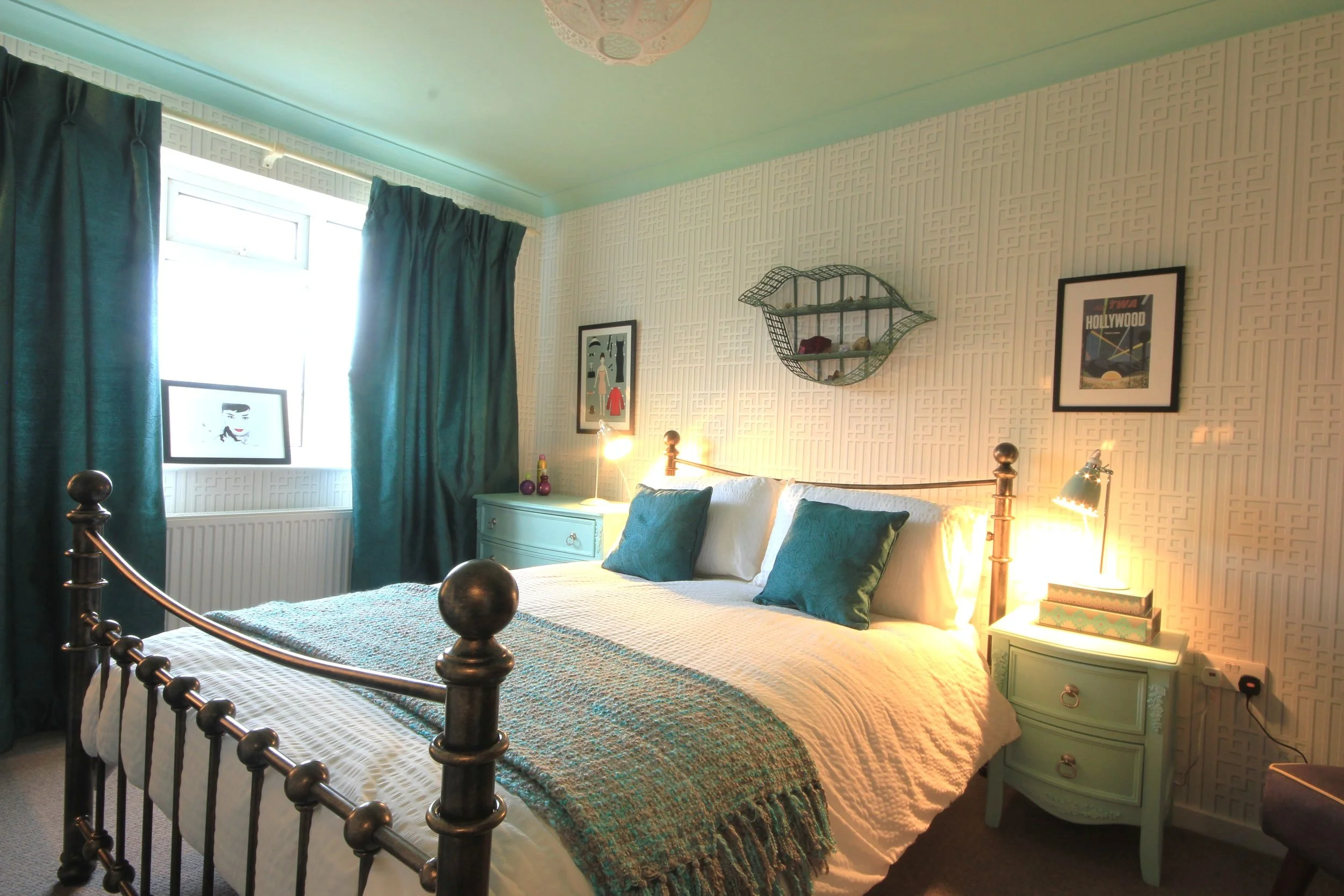 Bedroom with bronze metal bed in the centre. Light grey art deco wallpaper on all the walls. Mint green painted ceiling with bedside tables in the same colour