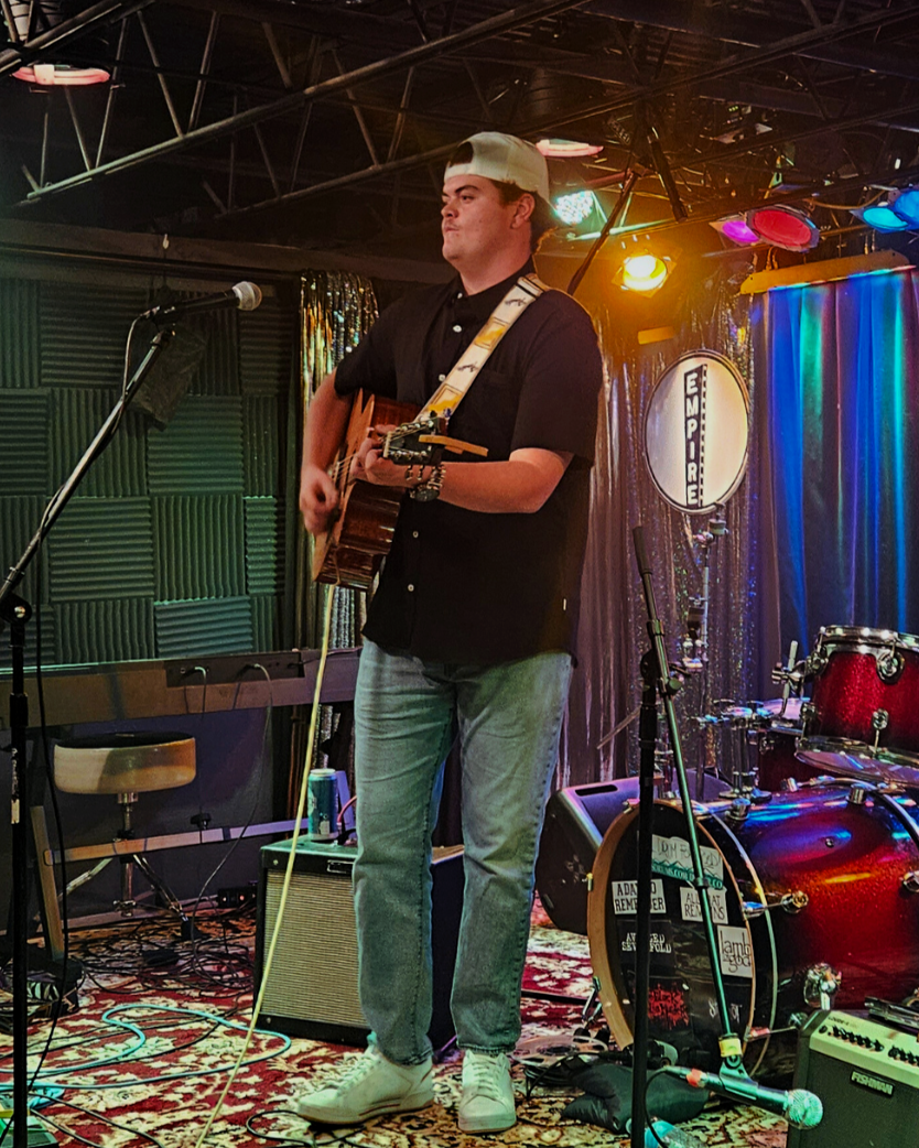 Young man wearing a backward white baseball cap, black shirt, and light jeans playing an acoustic guitar on stage with colorful stage lighting, drums, microphone, and sound equipment behind him.