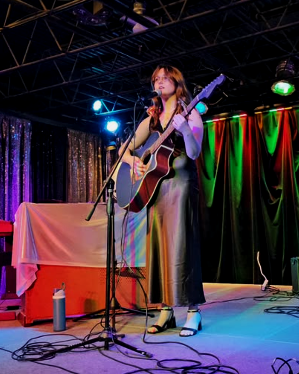 A woman with reddish-brown hair wearing a black dress and high-heeled sandals, playing an acoustic guitar and singing into a microphone on stage with vibrant colored lights and curtains in the background.