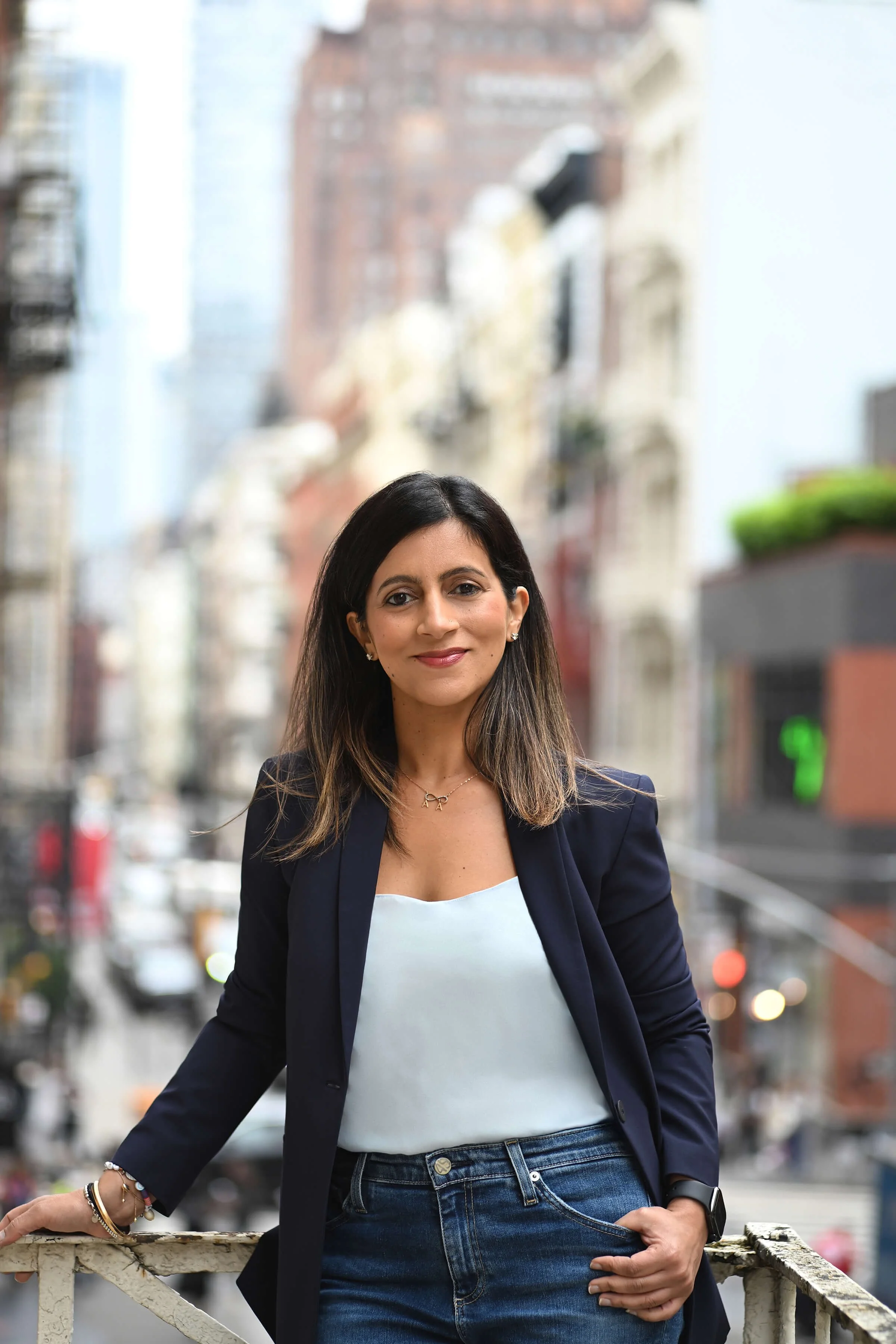 Mansi Narang standing on a fire escape in a blue blazer, silk top and denim jeans