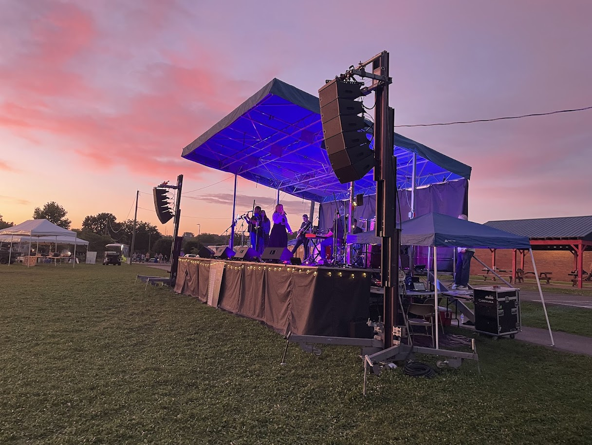 Outdoor concert stage with a band performing at sunset, featuring purple lighting, large speakers, and a grassy field.