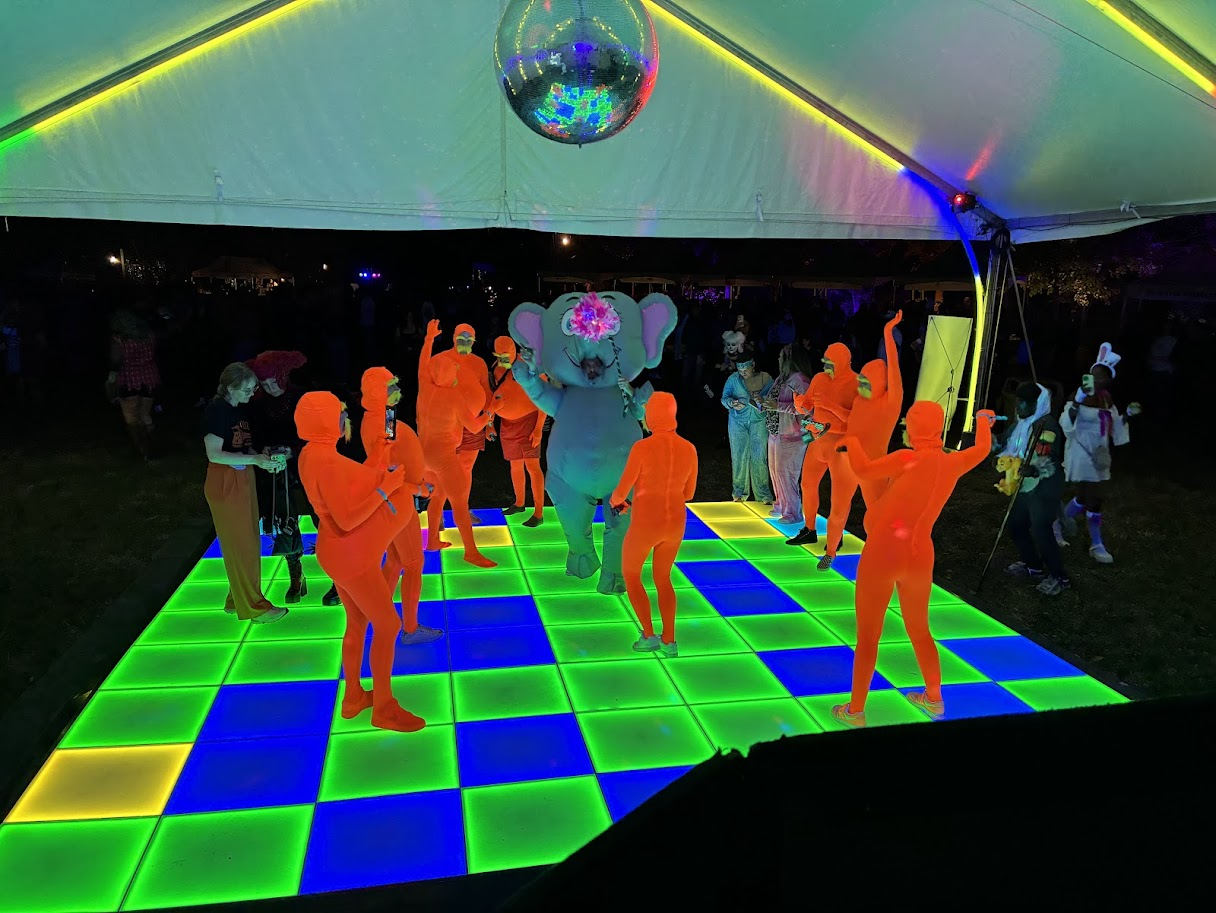 Guests dancing on a brightly lit, colorful illuminated dance floor at a nighttime party under a tent, with a person dressed in a large elephant costume with floral decorations on the head, and some people are dressed as animals.