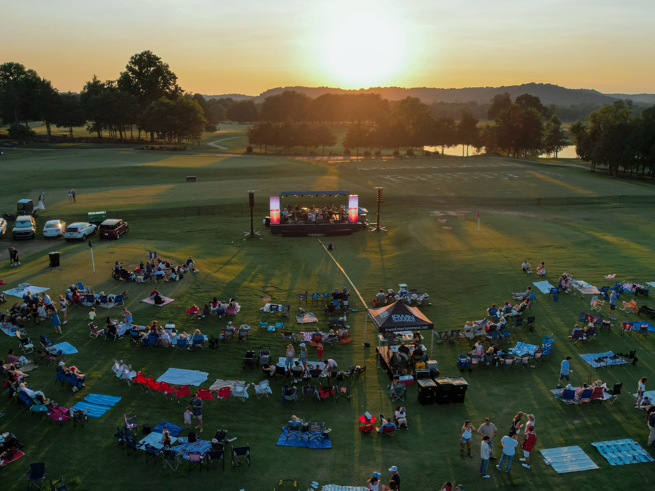 People gathered on a golf course lawn during sunset, attending an outdoor event with a stage, chairs, picnic blankets, and food trucks.