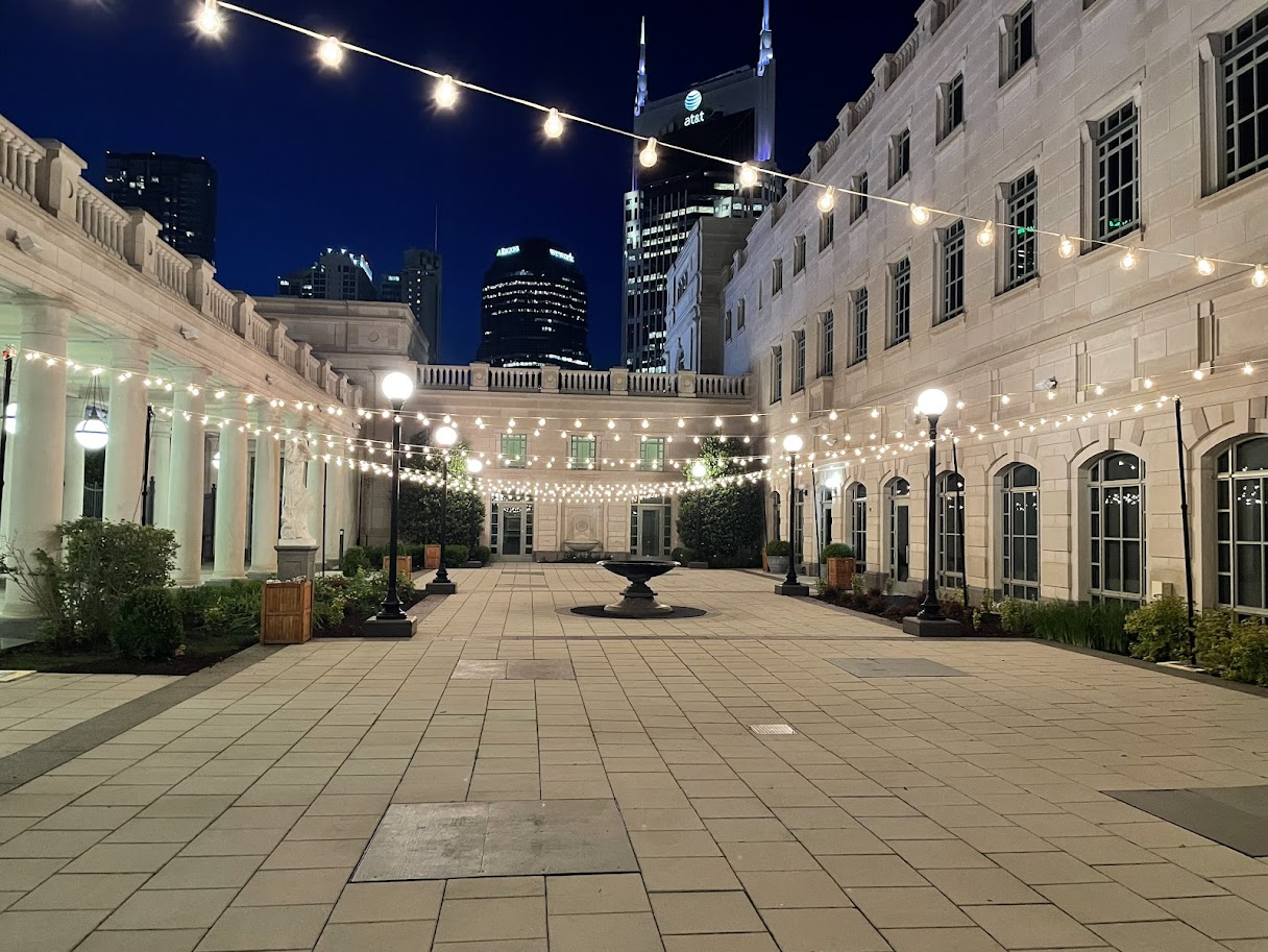 An outdoor courtyard at night with string lights hanging overhead, surrounded by white classical-style buildings with large windows, and a city skyline with tall illuminated skyscrapers in the background.