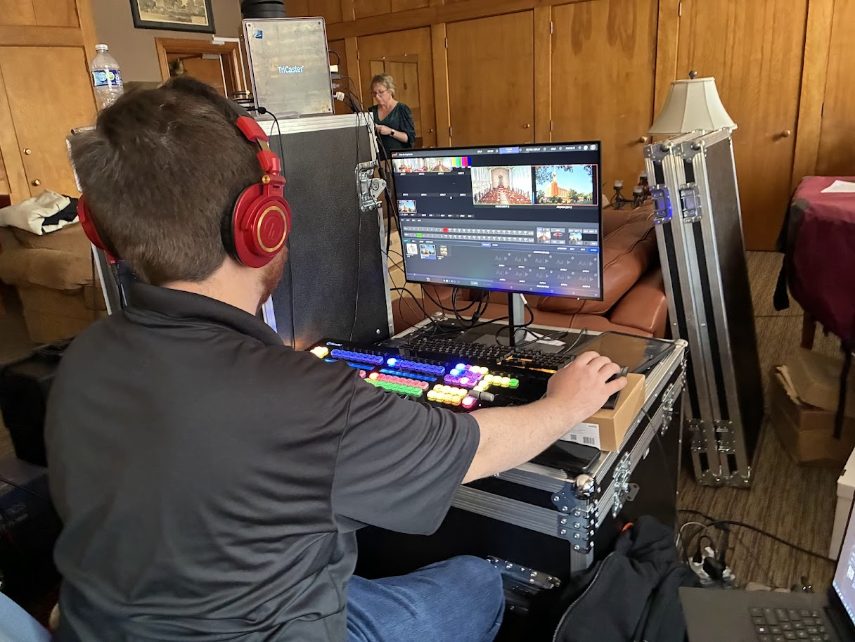 A man with red headphones operating a video editing machine at a desk, with a large monitor displaying video editing software. In the background, a woman is working on a computer.