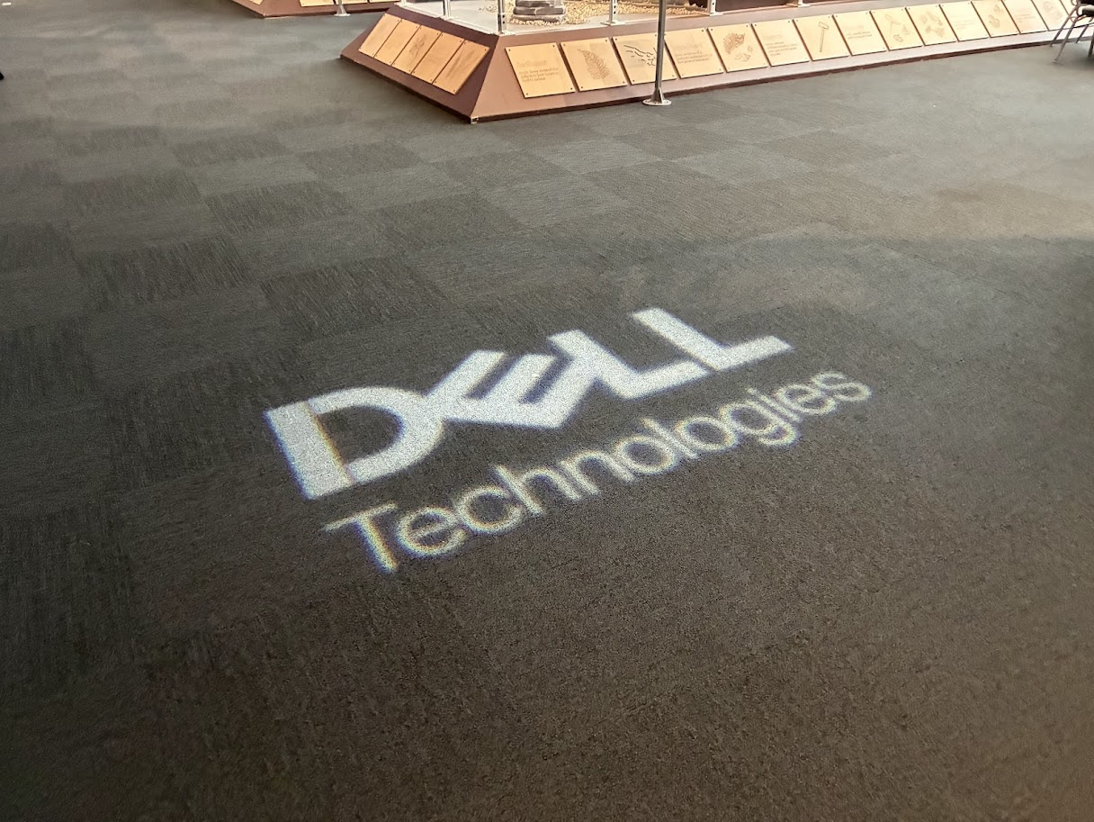 Projection of the Dell Technologies logo on a dark carpeted floor in an indoor setting, with a display case and informational plaques in the background.
