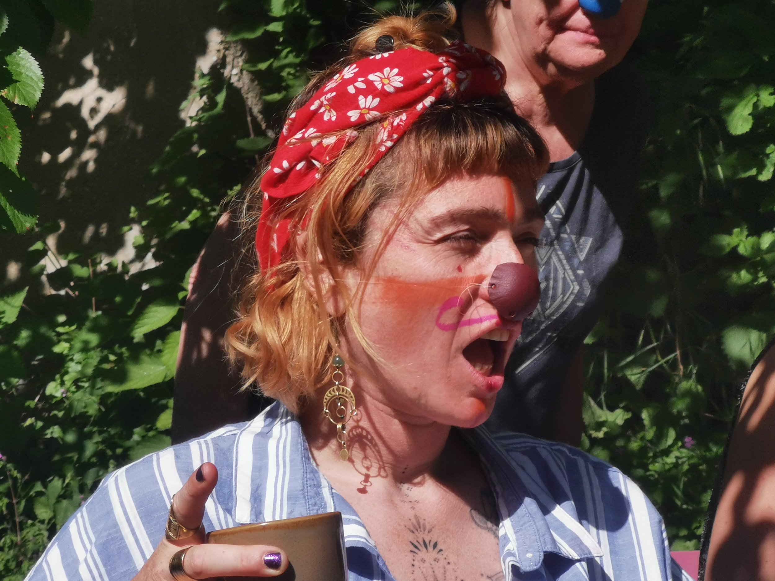 Une femme de profile, la bouche ouverte les yeux fermés, porte du maquillage et un nez de clown, elle a une frange et un bandana rouge sur la tête. Elle porte une chemise bleue rayée blanche et il y a de la verdure au second plan