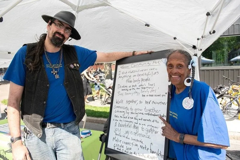 A tall white man in a hat and blue shirt stands next to a shorter black woman with braids. Both are smiling and displaying a community poem written collaboratively at a community event.