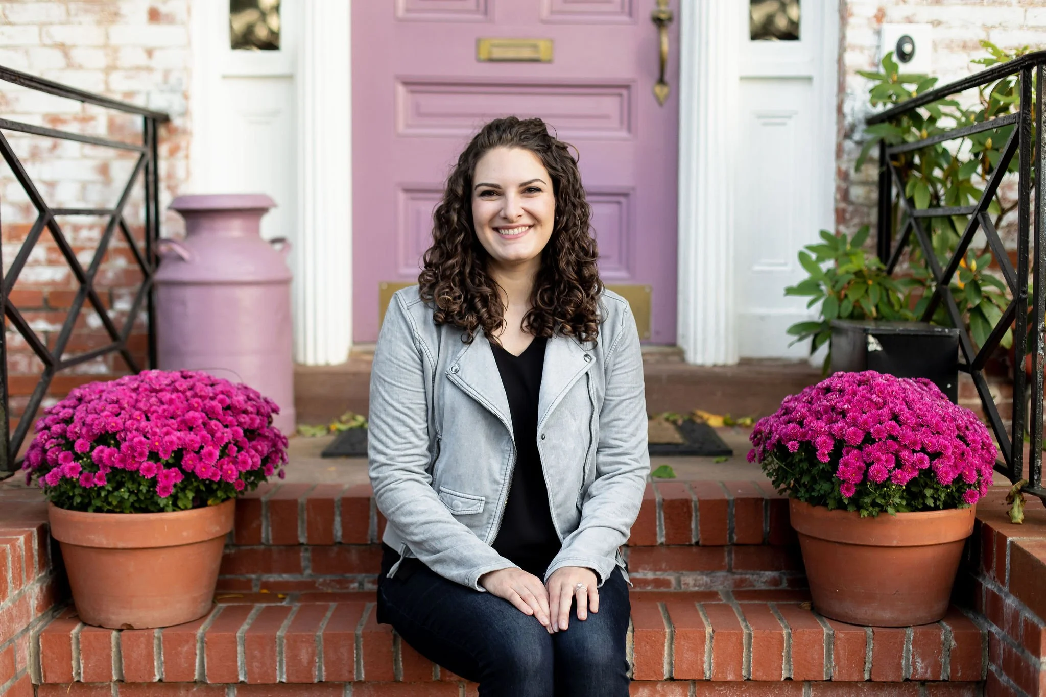 Image of Brittany Schnurr, LMFT, a therapist in Fairfax, VA sitting on brick steps of a historical home with a purple door with a warm smile and looking approachable. She is sitting beside two pots of fuscia mums.