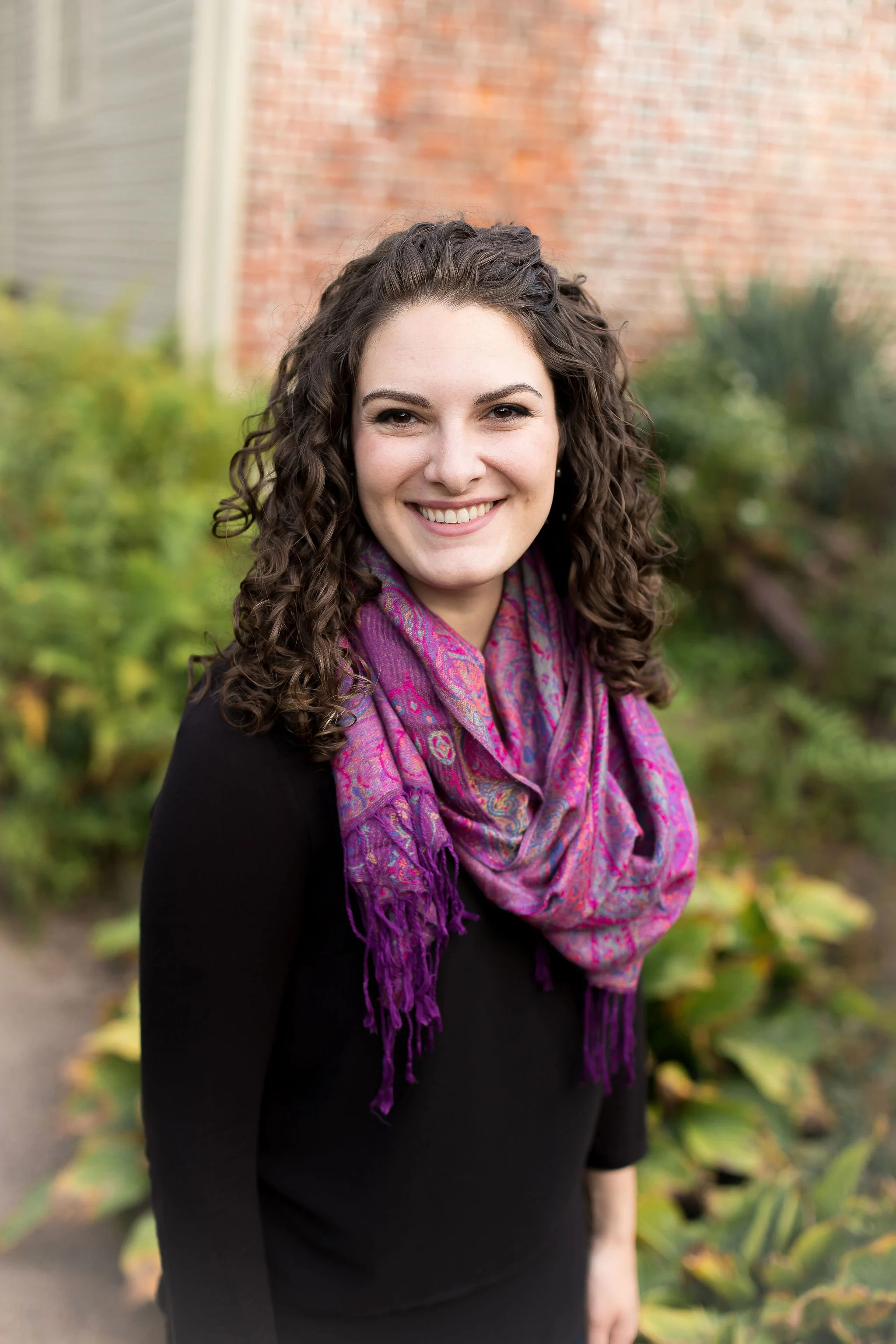 Image of Brittany Schnurr wearing a purple scarf and standing outside by a historical brick building and spring foliage. Brittany Schnurr, LMFT is a therapist serving women in Fairfax and all over Virginia and Connecticut.