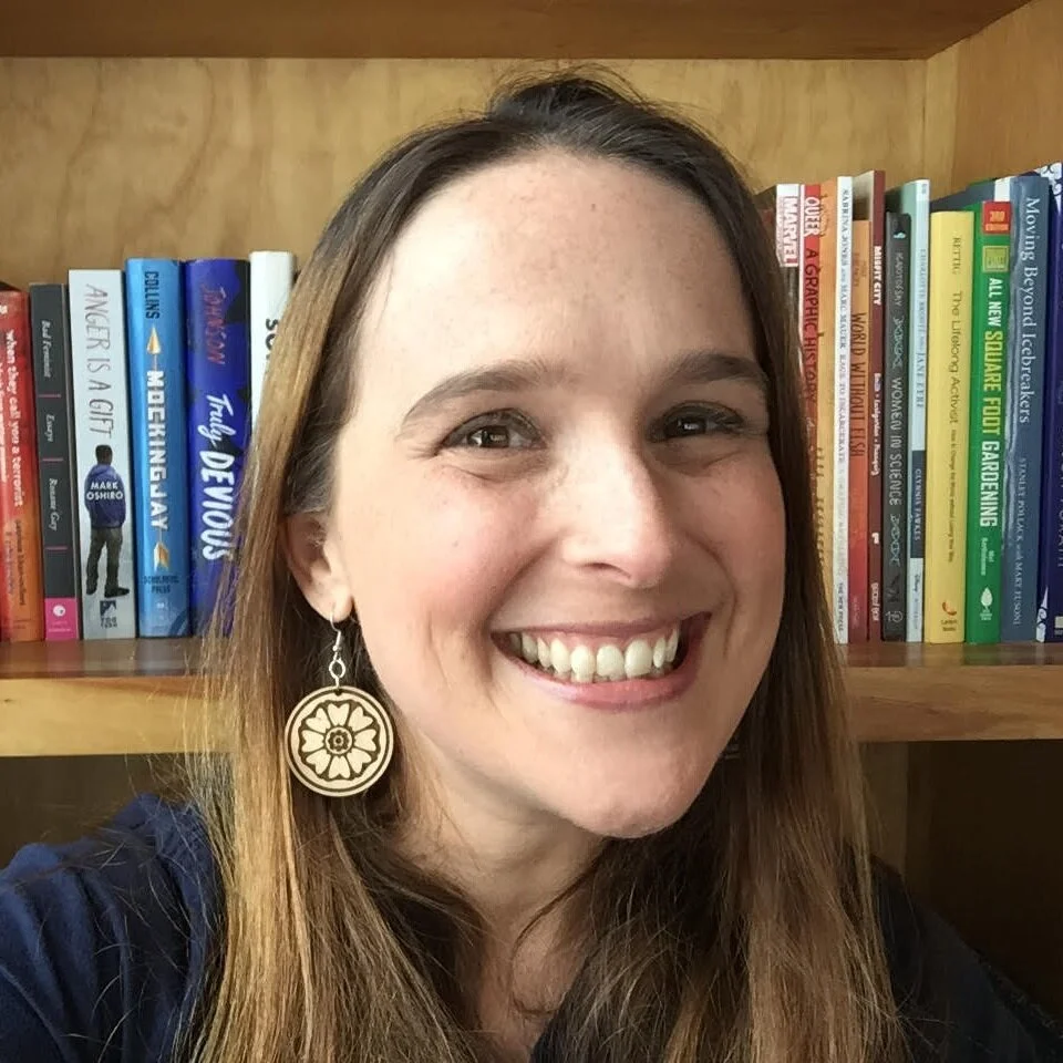 Katie Bowers, a white woman with straight light brown hair, smiles at you. She is standing in front of her bookshelf and wearing an awesome pair of large, flowery earrings.