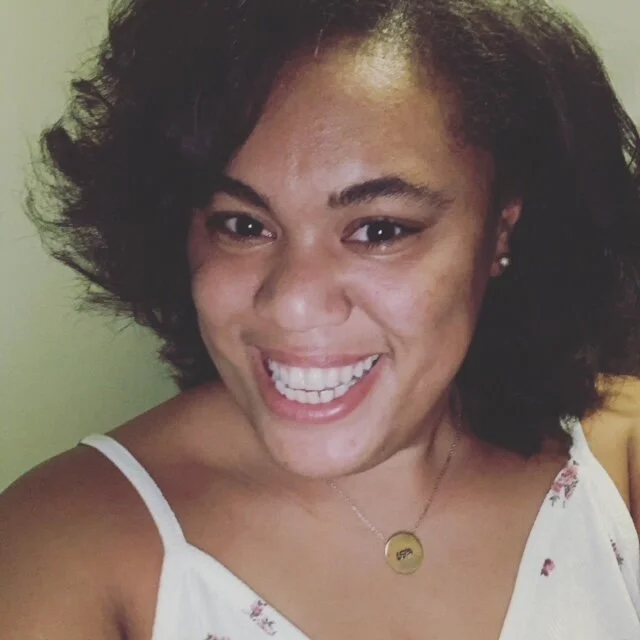 K. Alexandra Mills, a Black woman with wavy brown hair, smiles at you. She is wearing a white shirt and a gold necklace.