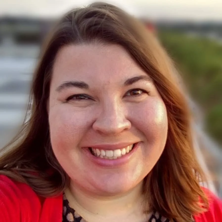 Maggie Brevig, a white woman with straight light brown hair, smiles at you. She is wearing a black and pink shirt and a red cardigan.