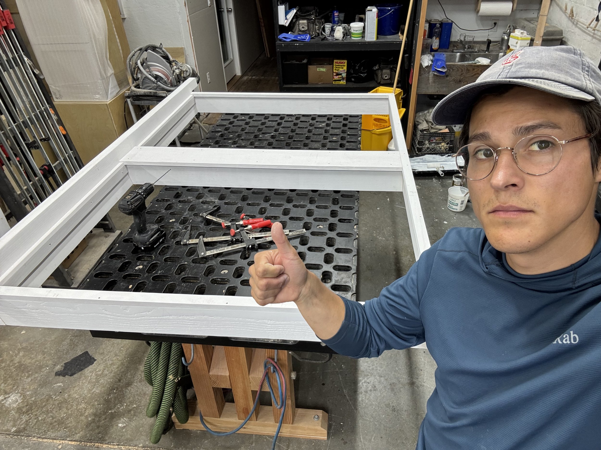 A man giving a thumbs up in front of a window frame. The window frame is primed and the man is wearing gold glasses and a baseball hat