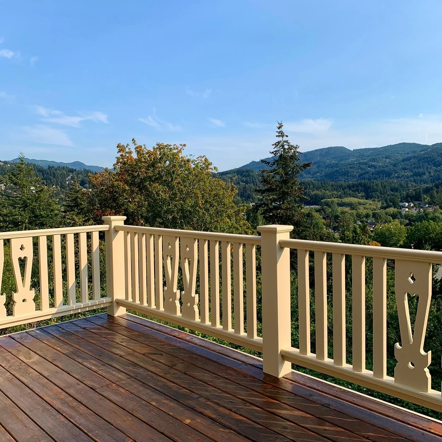 Wood deck with a cream-colored railing featuring decorative cutout balusters, overlooking hills under a blue sky.
