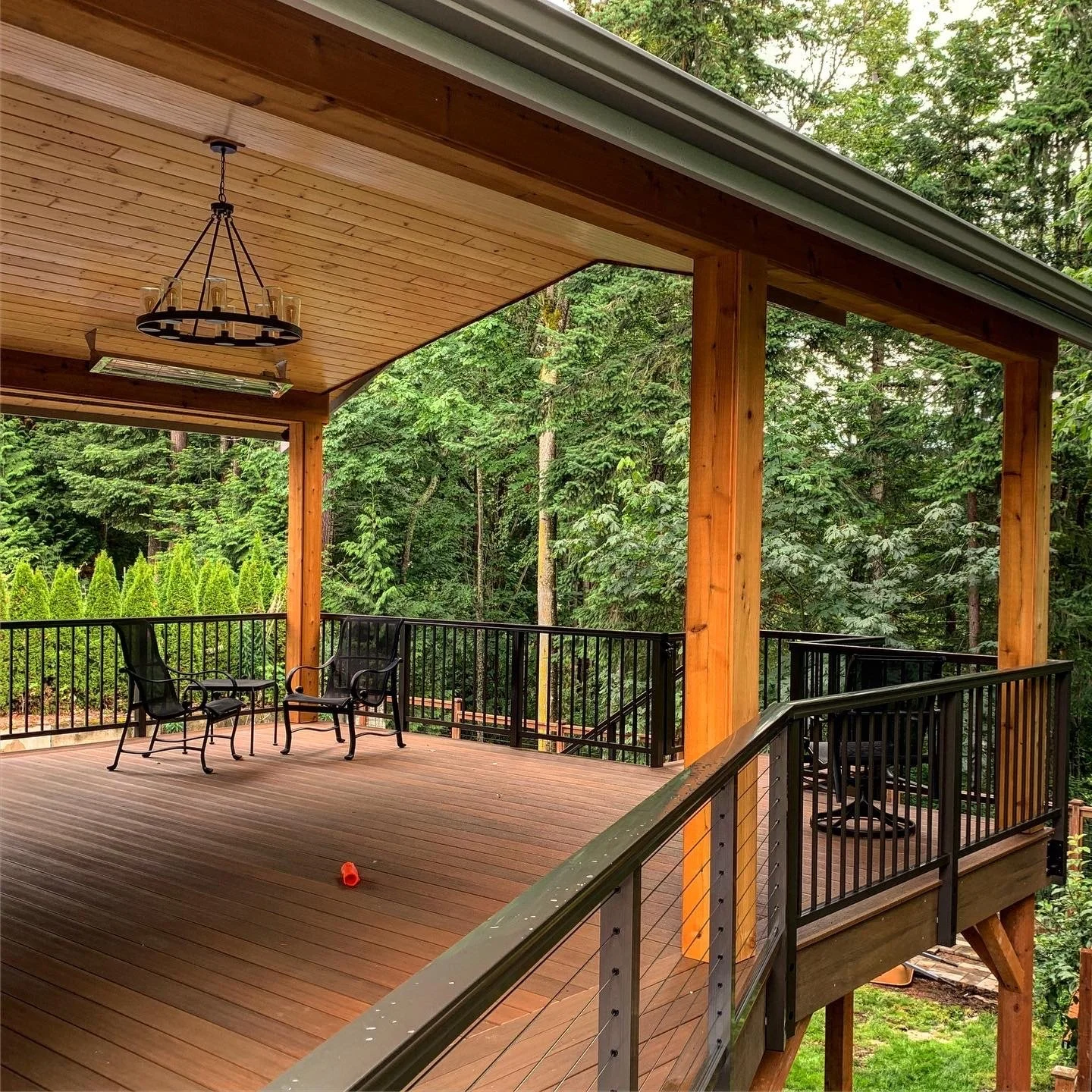 Covered deck with a wood plank ceiling, black metal railing, hanging black chandelier, and patio chairs, surrounded by trees.