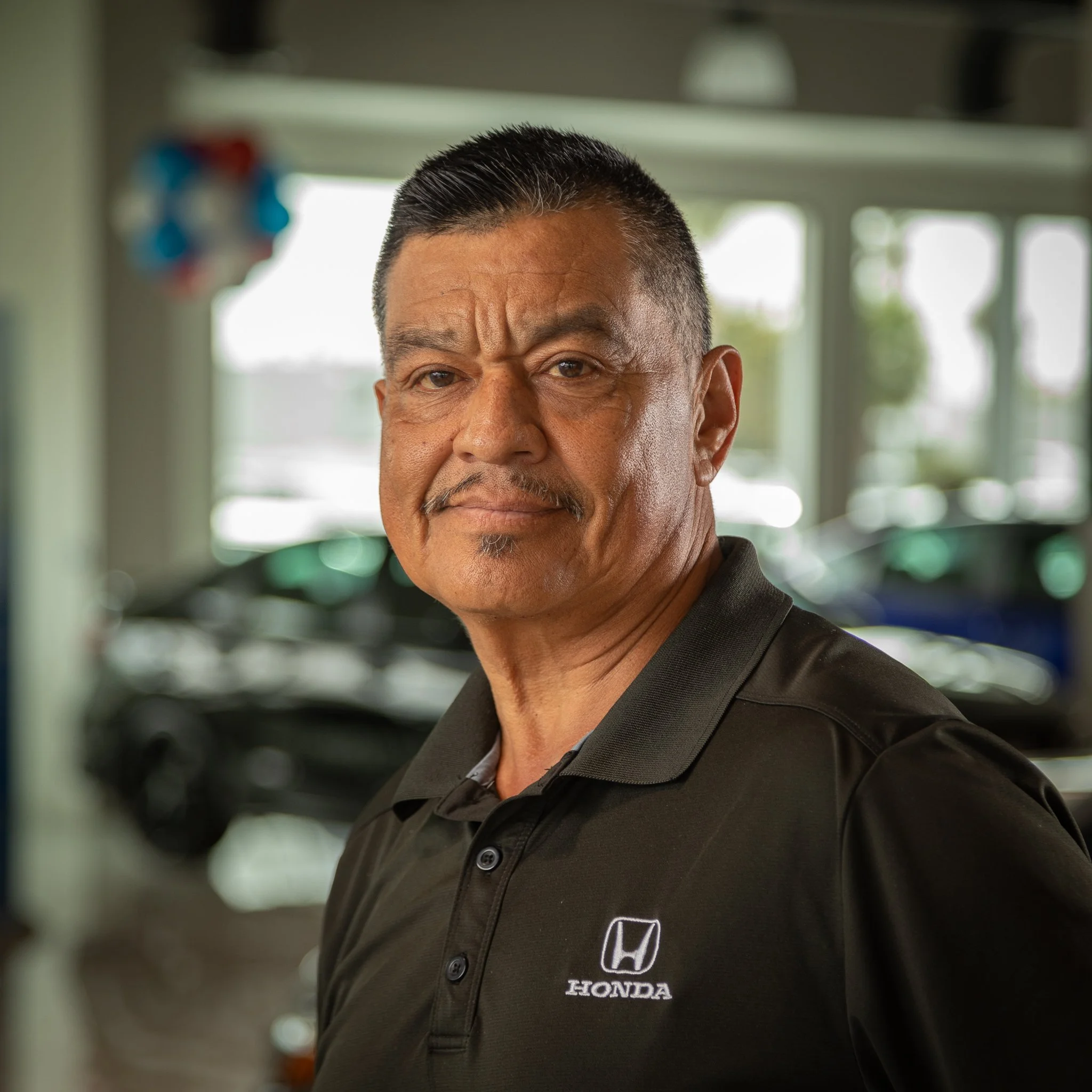 A man with short dark hair, a mustache, and a goatee standing in a car dealership, wearing a black Honda polo shirt, with cars visible in the background.
