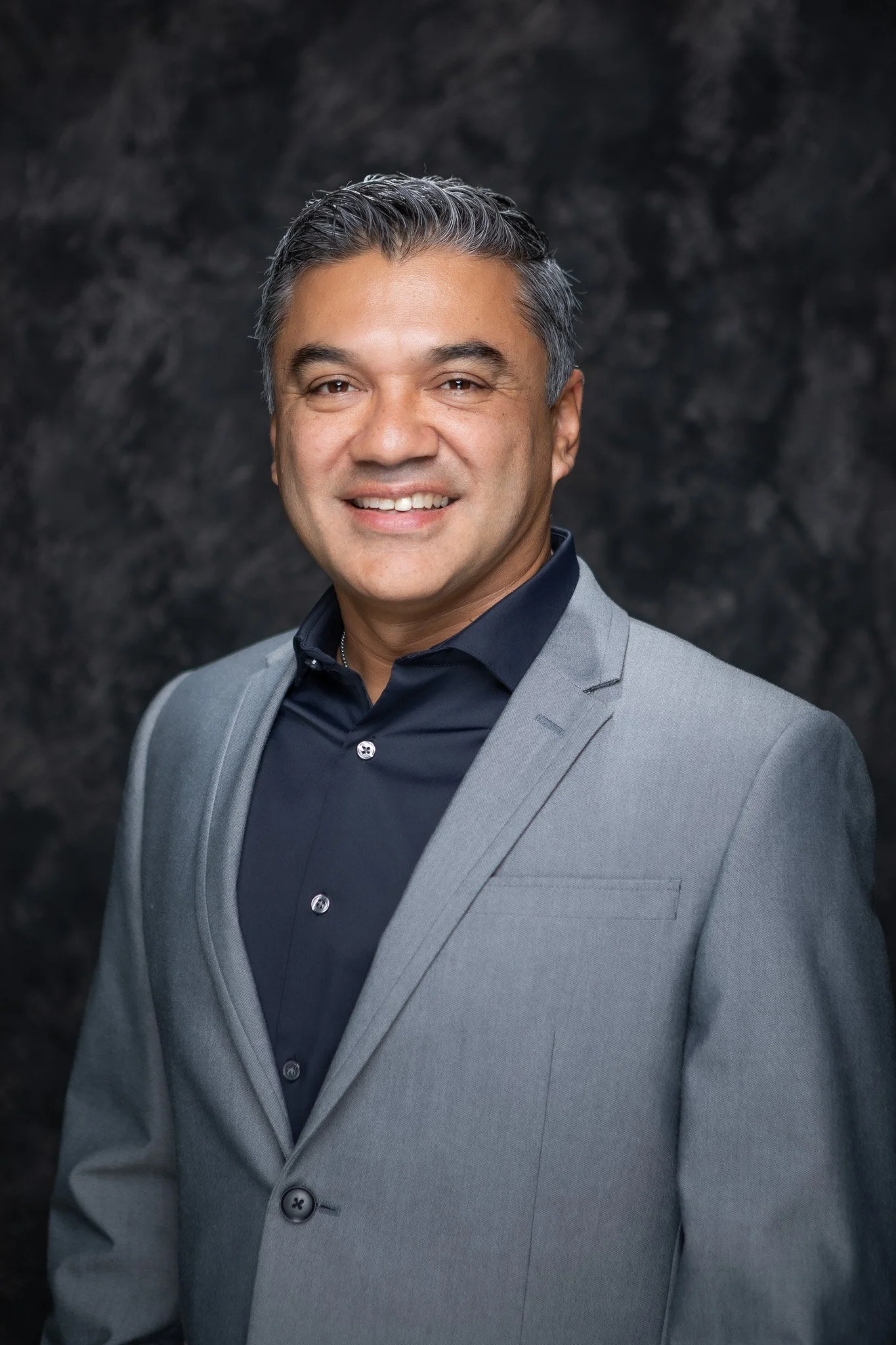 Professional headshot of a smiling man in a gray suit and dark blue shirt against a black background.