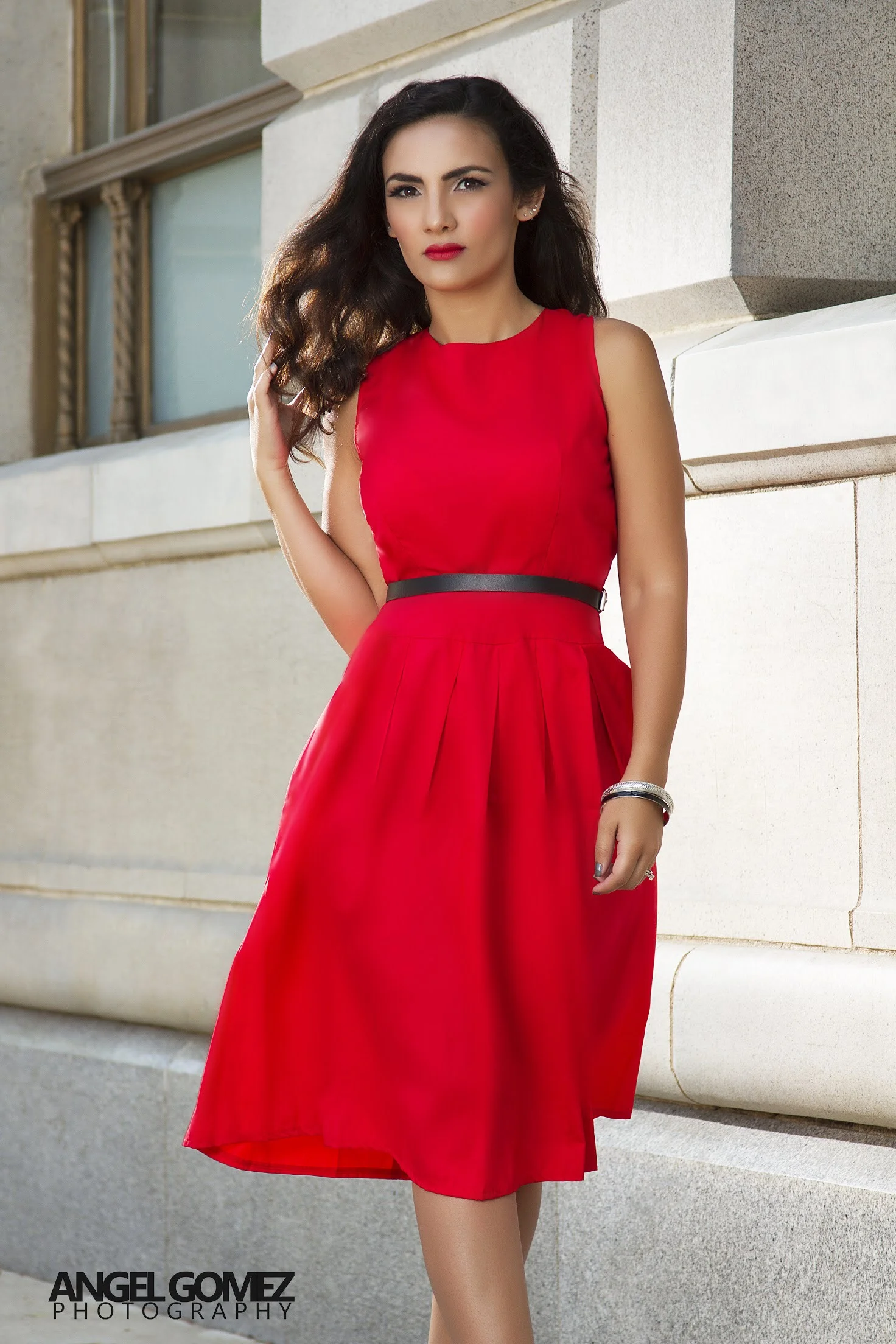 A woman with dark wavy hair wearing a sleeveless red dress and a black belt, standing against a stone building.